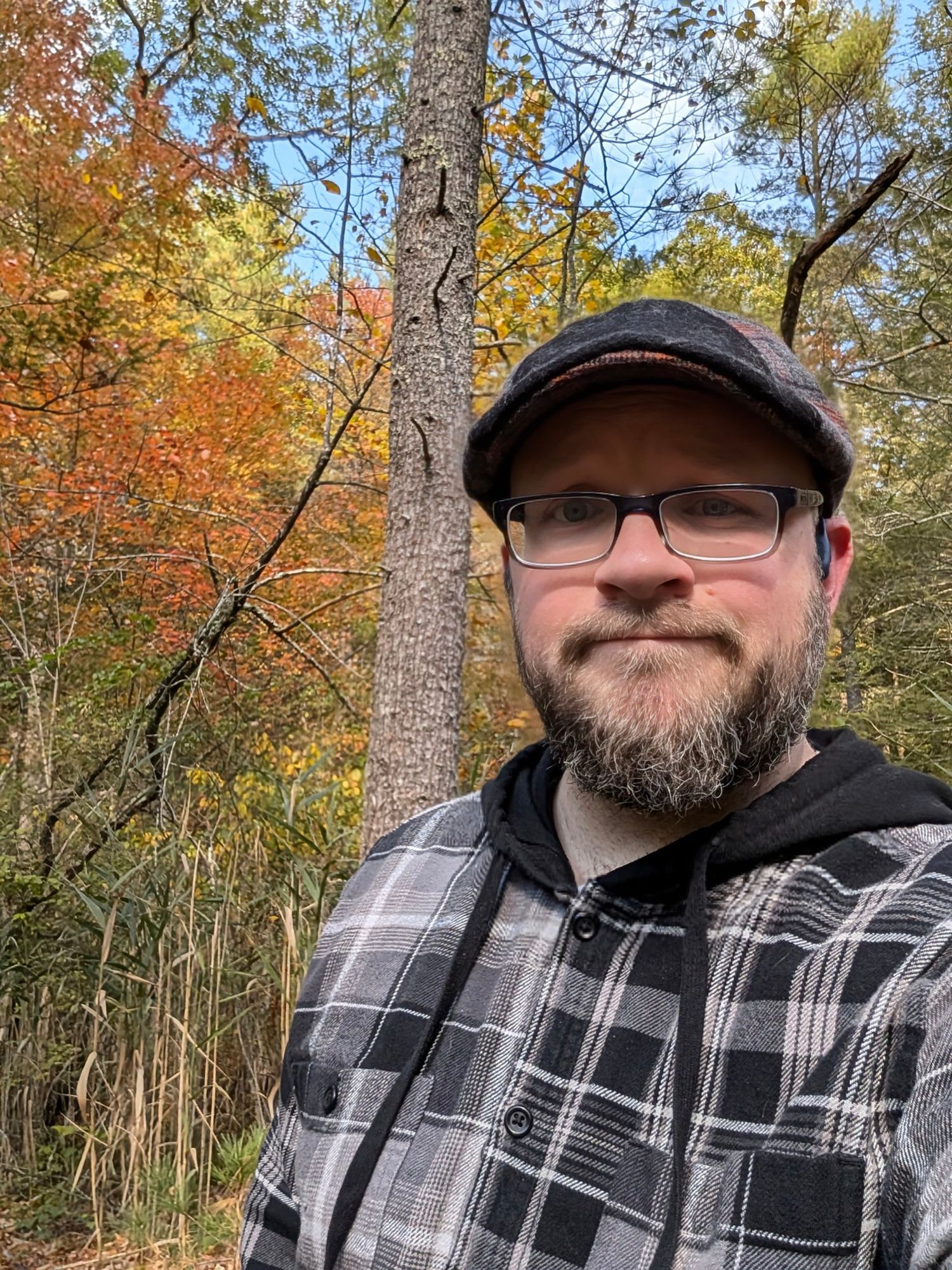 Selfie of a bearded man in flannel and a scally cap in the woods with yellow and orange foliage