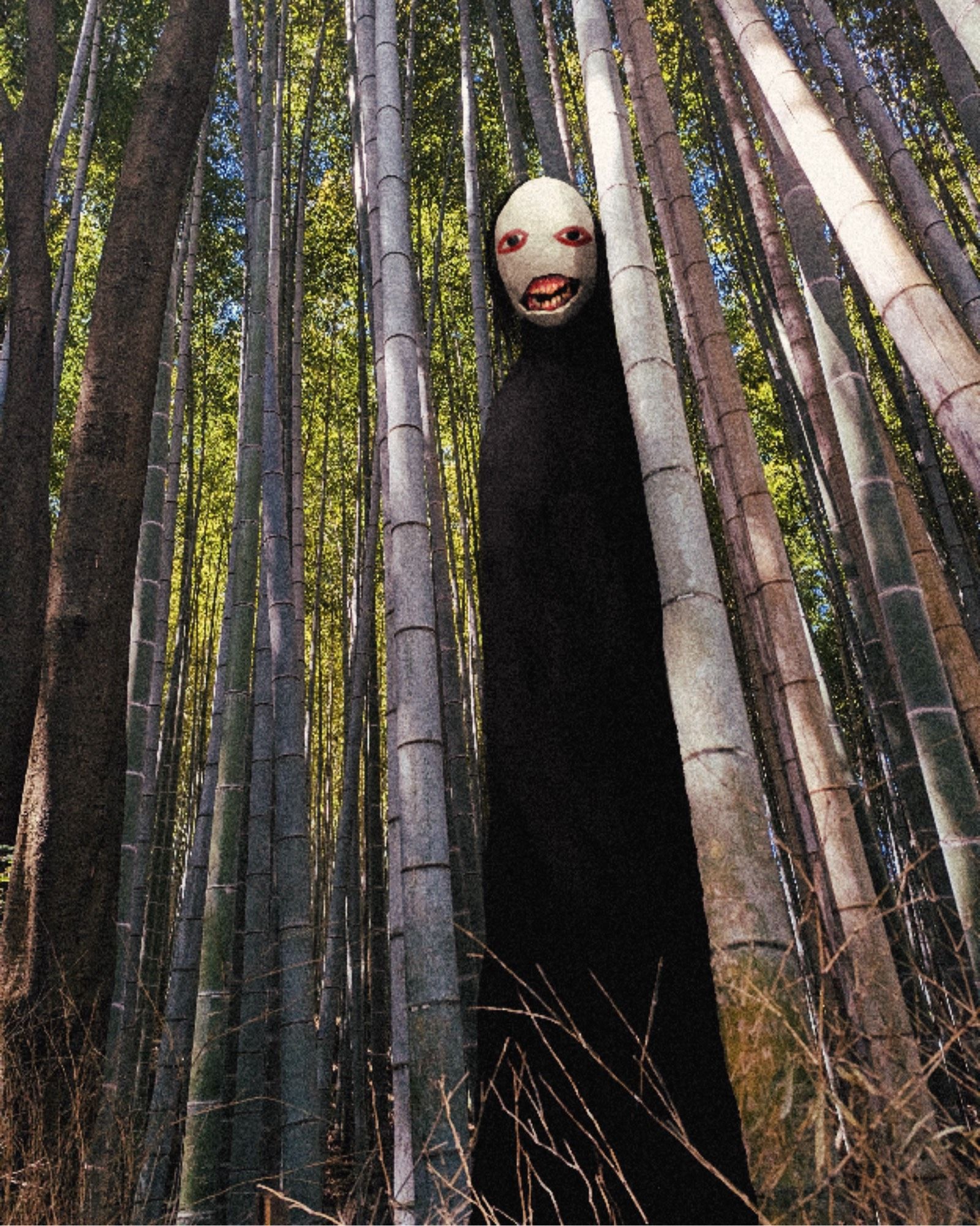 A tall figure in a black cloak lurks among bamboo trees. It wears a white mask with painted eyes. The mask has a crack through which rotten teeth and gums can be seen.