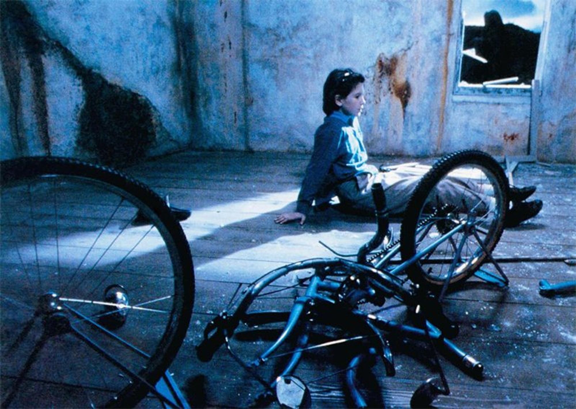 a young girl sits in a decrepit room, next to a broken bicycle.