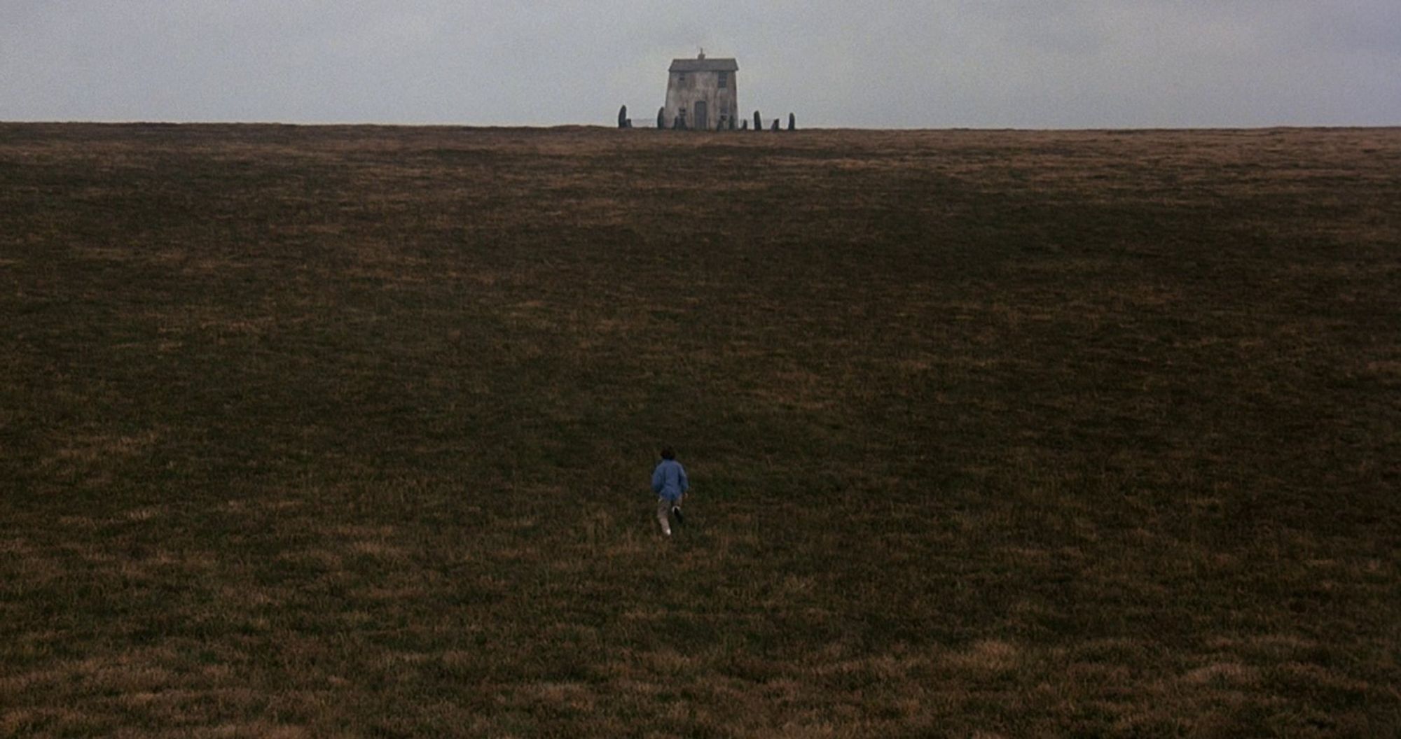 A young girl runs through an empty field. In the distance, a warped grey house stands surrounded by stones and a fence.