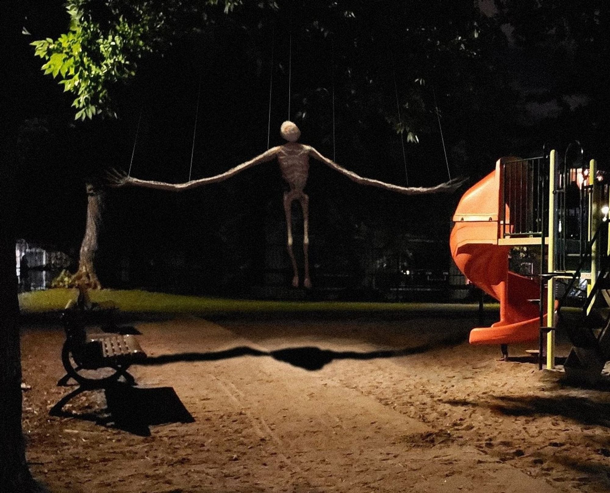 a faceless figure hung from strings in a playground, its arms are exceptionally long