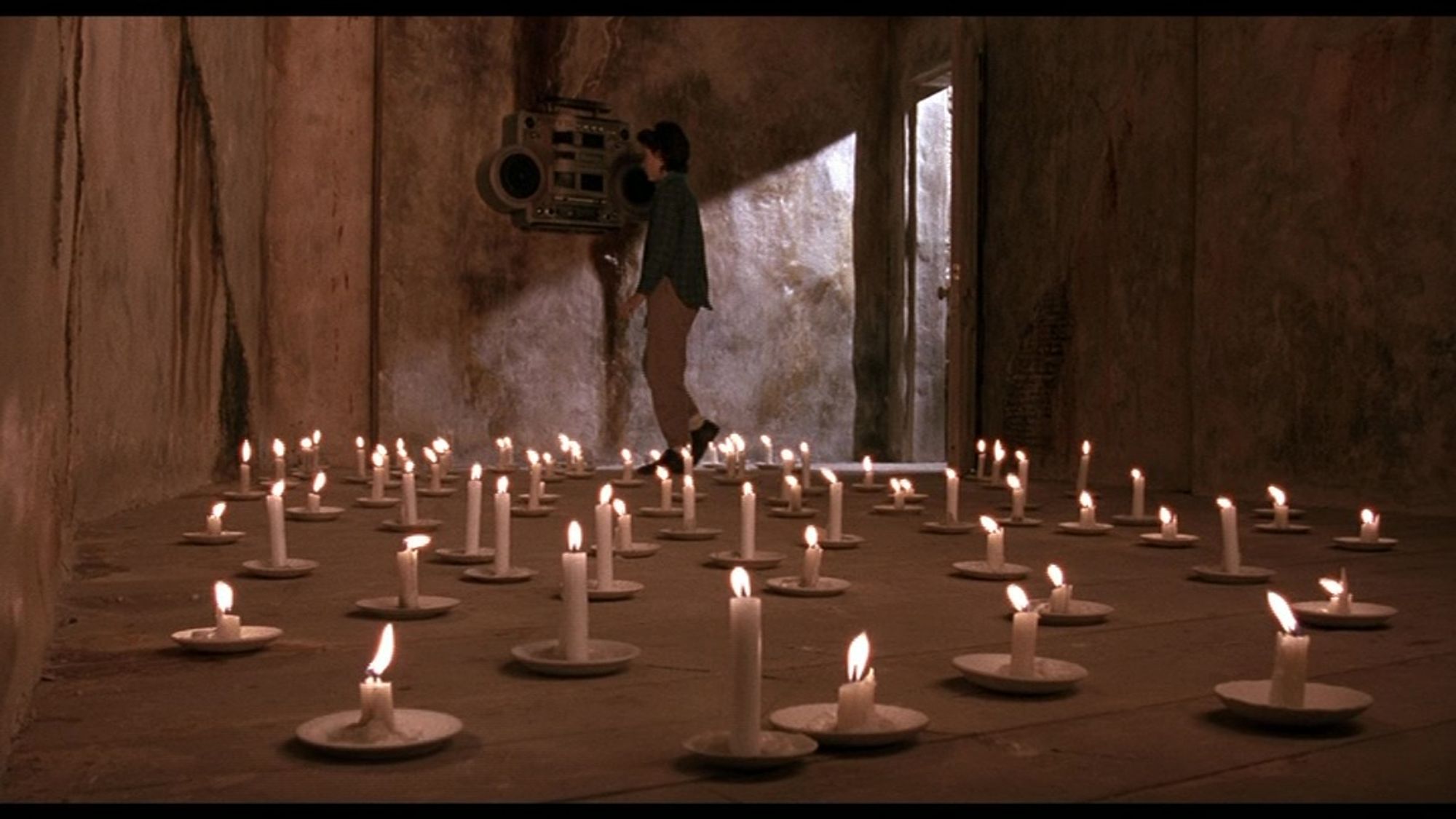 A young girl inspects a strange music player on the wall of an empty room. the floor is covered by lit candles.