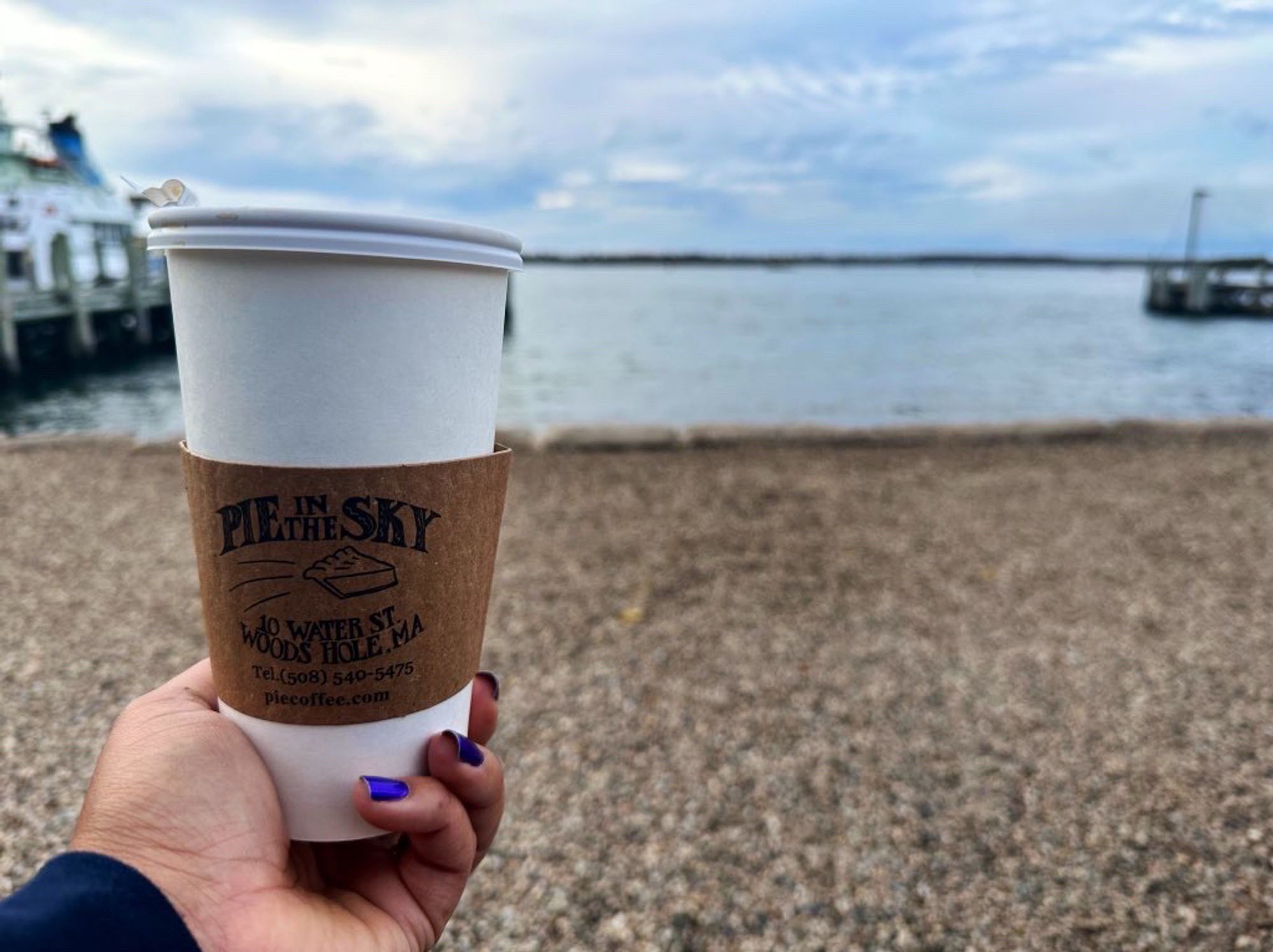 A woman holding a pie in the sky coffee cup in front of the waterfront in woods hole.