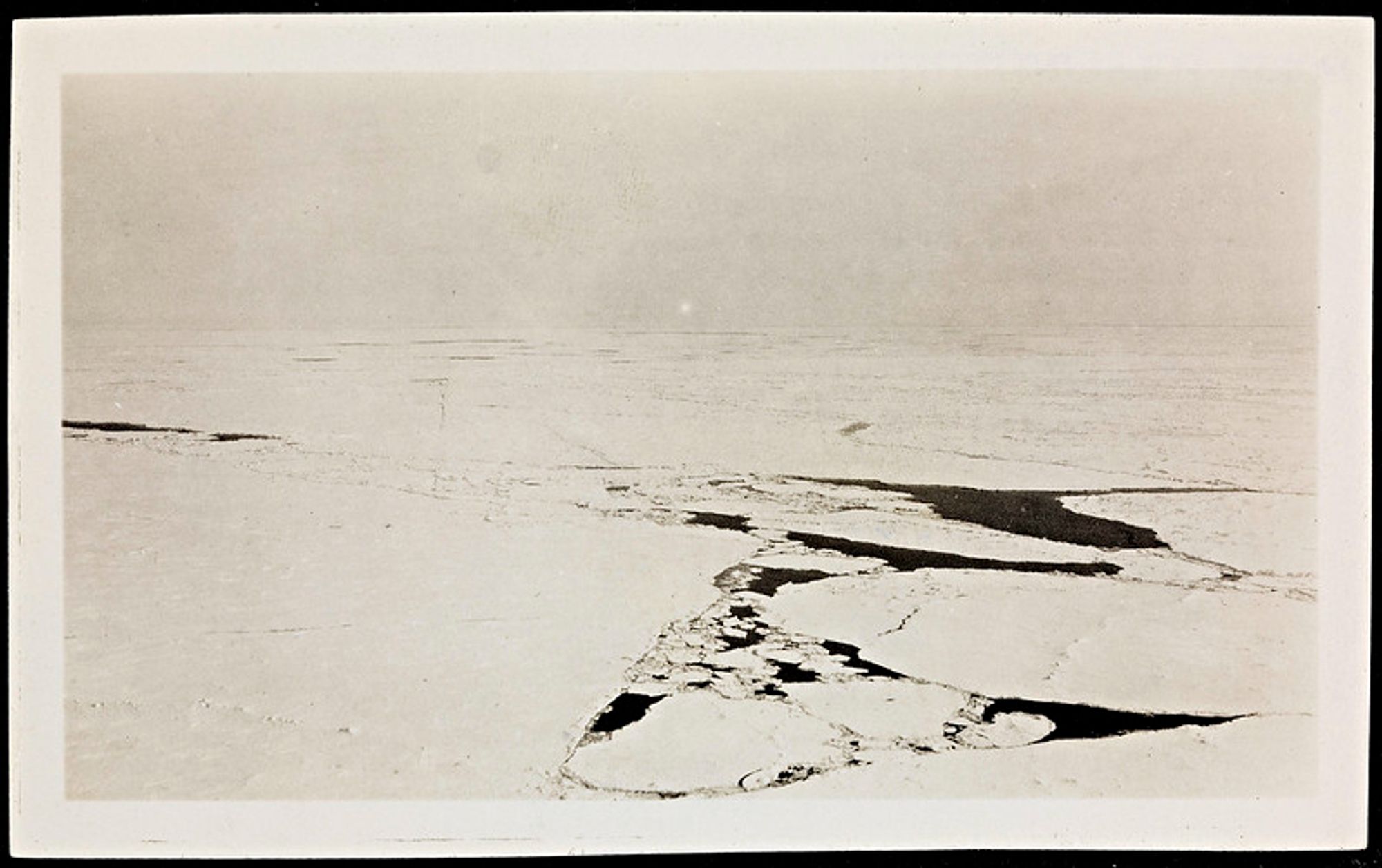 a grey sky over a slightly lighter ice field which has a few cracks in it revealing the darker waters below.