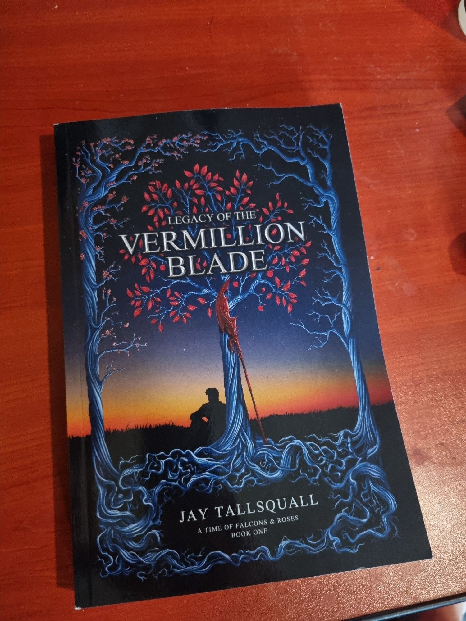 This picture shows the book, Legacy of thy Vermillion Blade, by Jay Tallsquall.
The cover art shows a man sitting under a tree, the wood the tree is silver, the leaves are red, there's a darker red halberd in front of the trunk of the tree