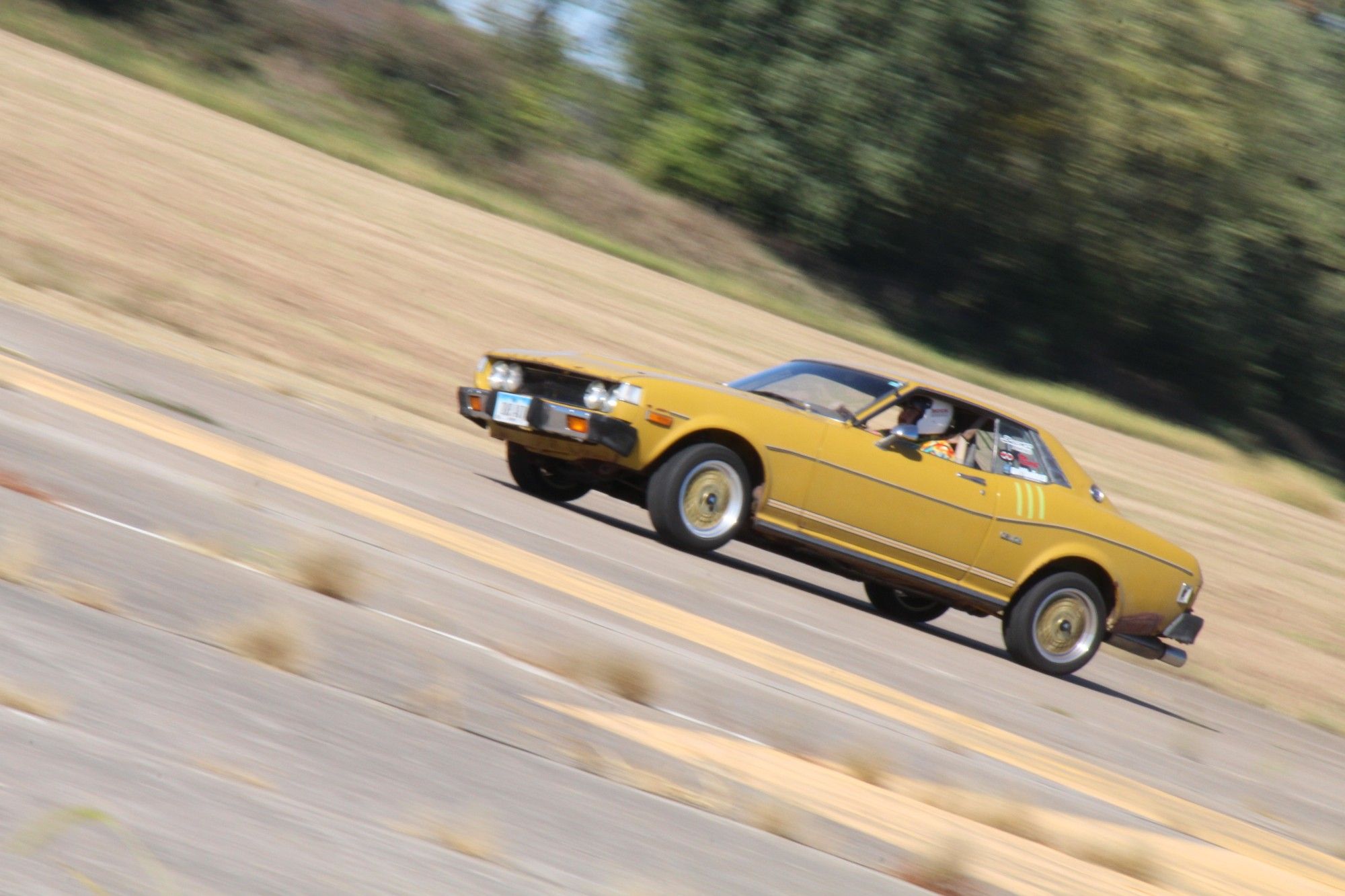 A mustard yellow 70s Toyota Celica racing on course.