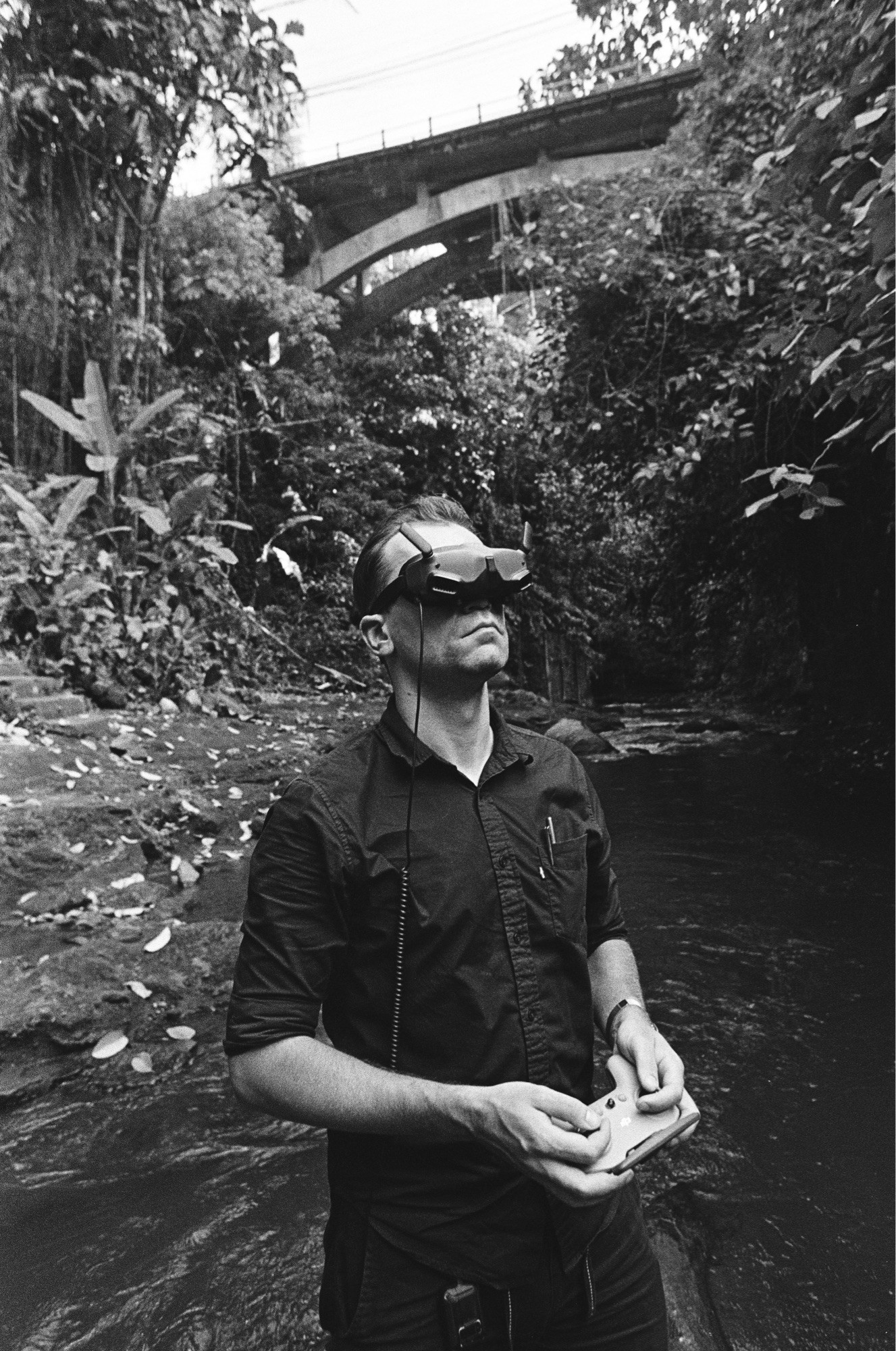 Man controlling a drone. On the mans face there are fpv googles. Background is jungle bridge and a river.