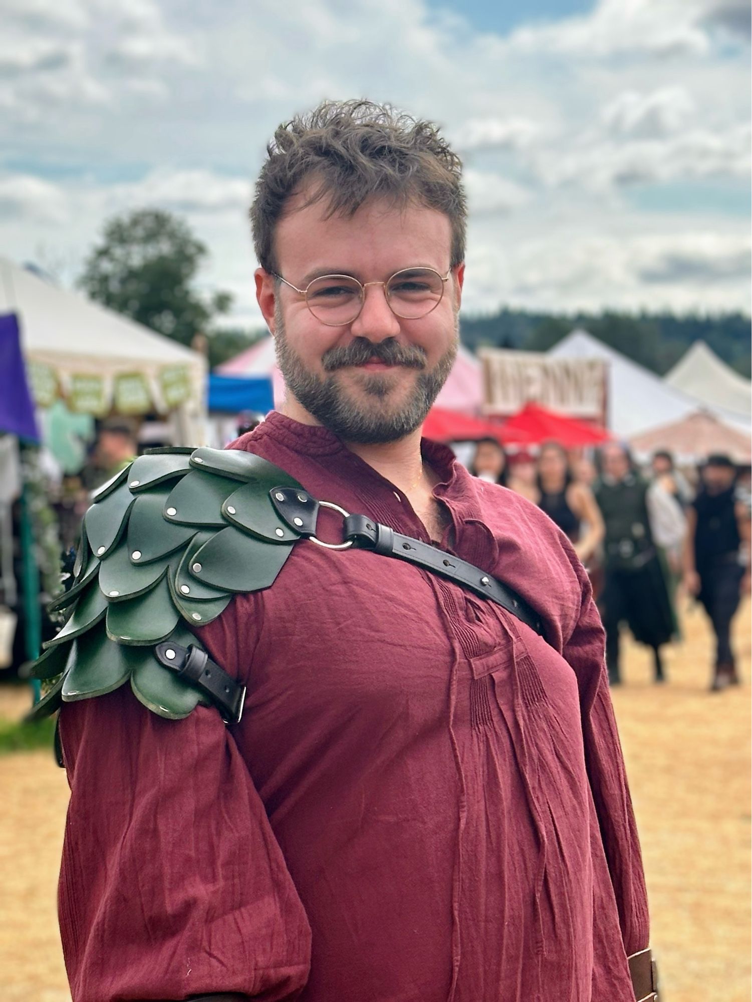 Gav stands, facing the camera in a maroon cotton Renaissance Faire-style shirt. The shoulder closest to the camera has a leather pauldron of dark green scales. It’s a beautiful day at the Renn Faire, and they’re giving the camera a tight grin with a squint.