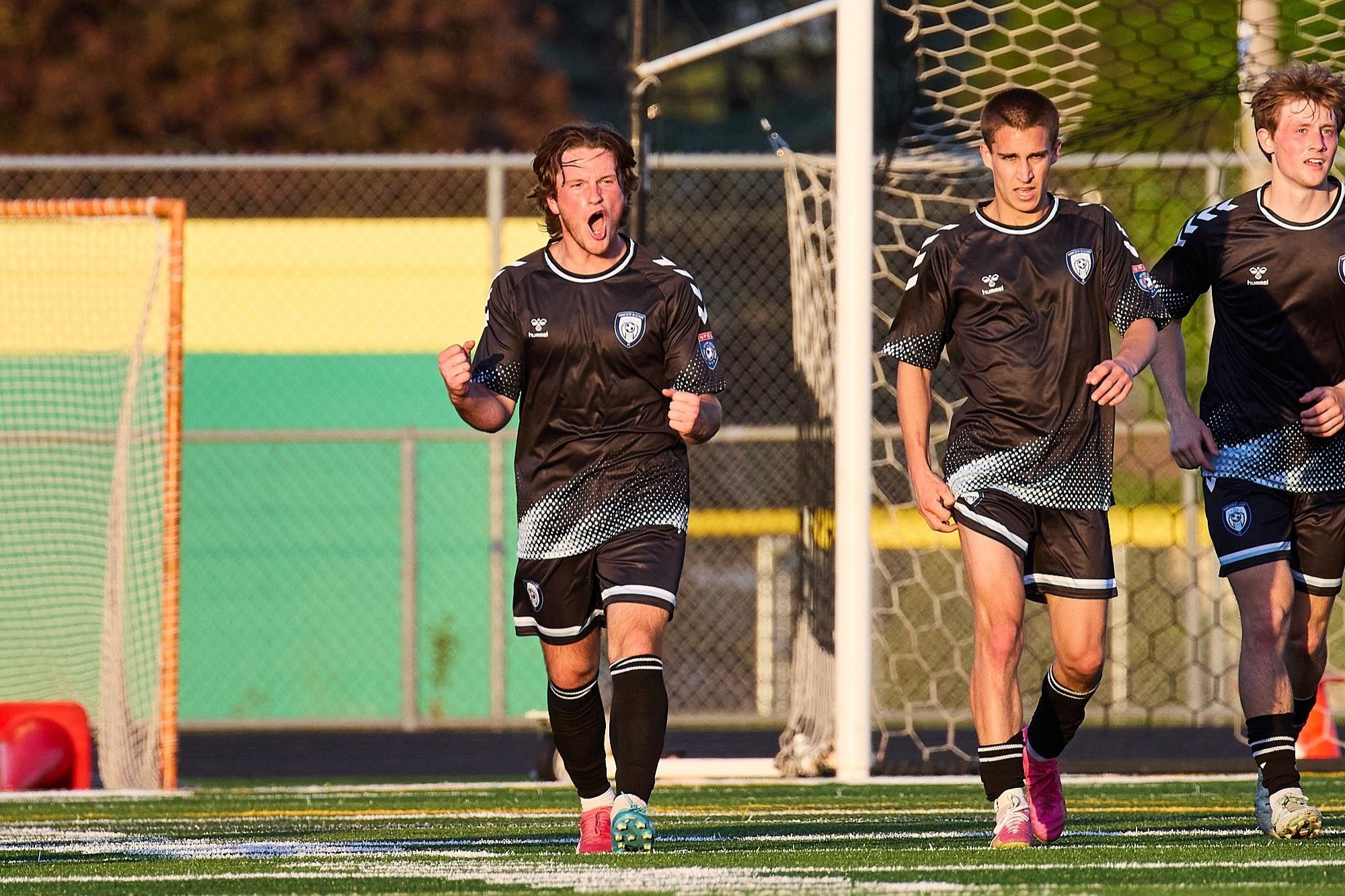 A trio of soccer players in black shirts and shorts celebrate a goal. The player on the left of frame has his mouth open in a bellow.