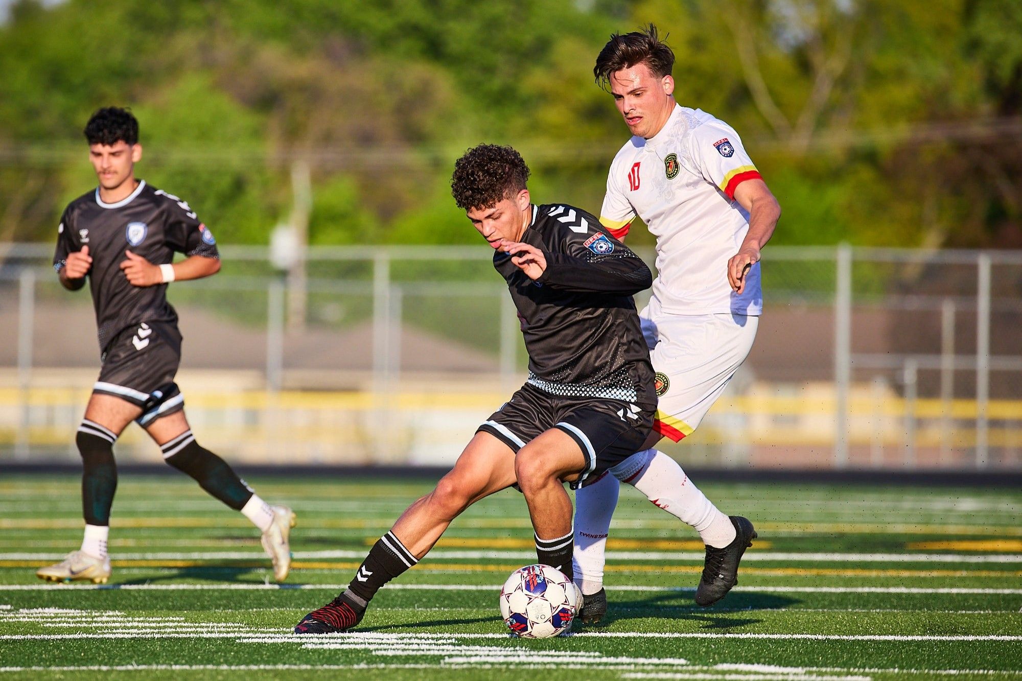 Player in black shields the ball from a tackle from a player in white.