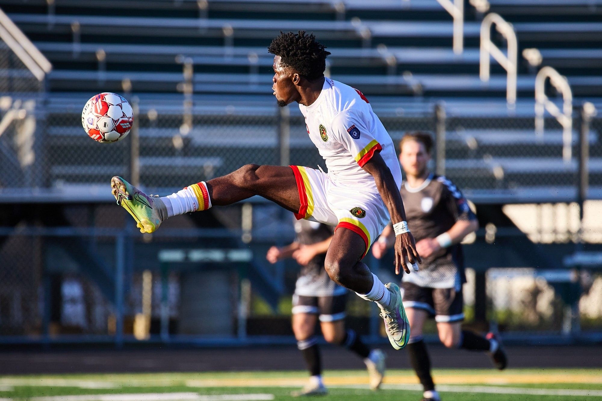 A white kitted soccer player is pictured up in the air, with the ball just to the left of his boot