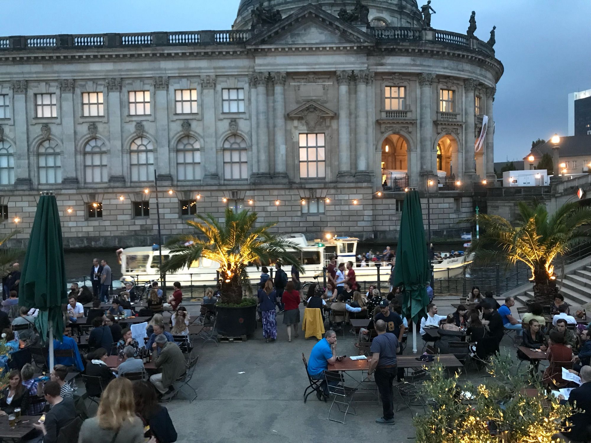 Vordergrund Menschen an Tischen sitzend. Dahinter Tanzfläche mit beleuchteten Palmen und Menschen. Dahinter Spree mit Schiff. Und dahinter Museumsbau.