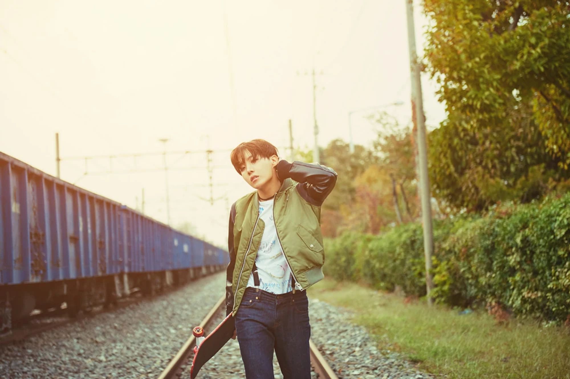 J-Hope in 2015 from the Most Beautiful Moment in Life Part 2. He's walking on traintracks while wearing a green and black jacket, and blue jeans. He's holding a skateboard. The setting sun washes the image in a soft orange