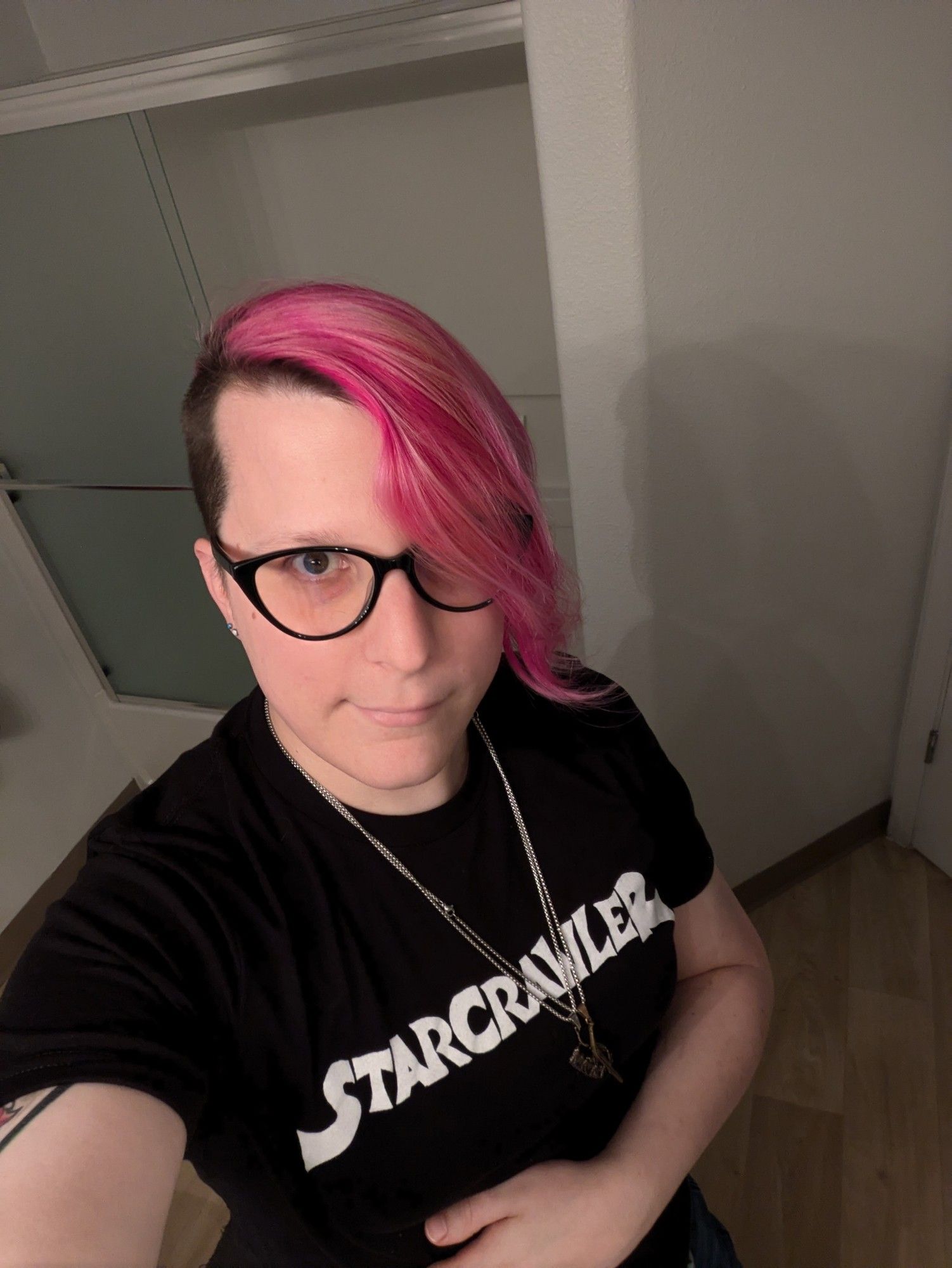 lowercase judy with fading magenta hair swept across one eye. she is wearing cat-eye glasses and a black shirt that says "STARCRAWLER" on it. she is doing a stereotypical trans selfie smirk