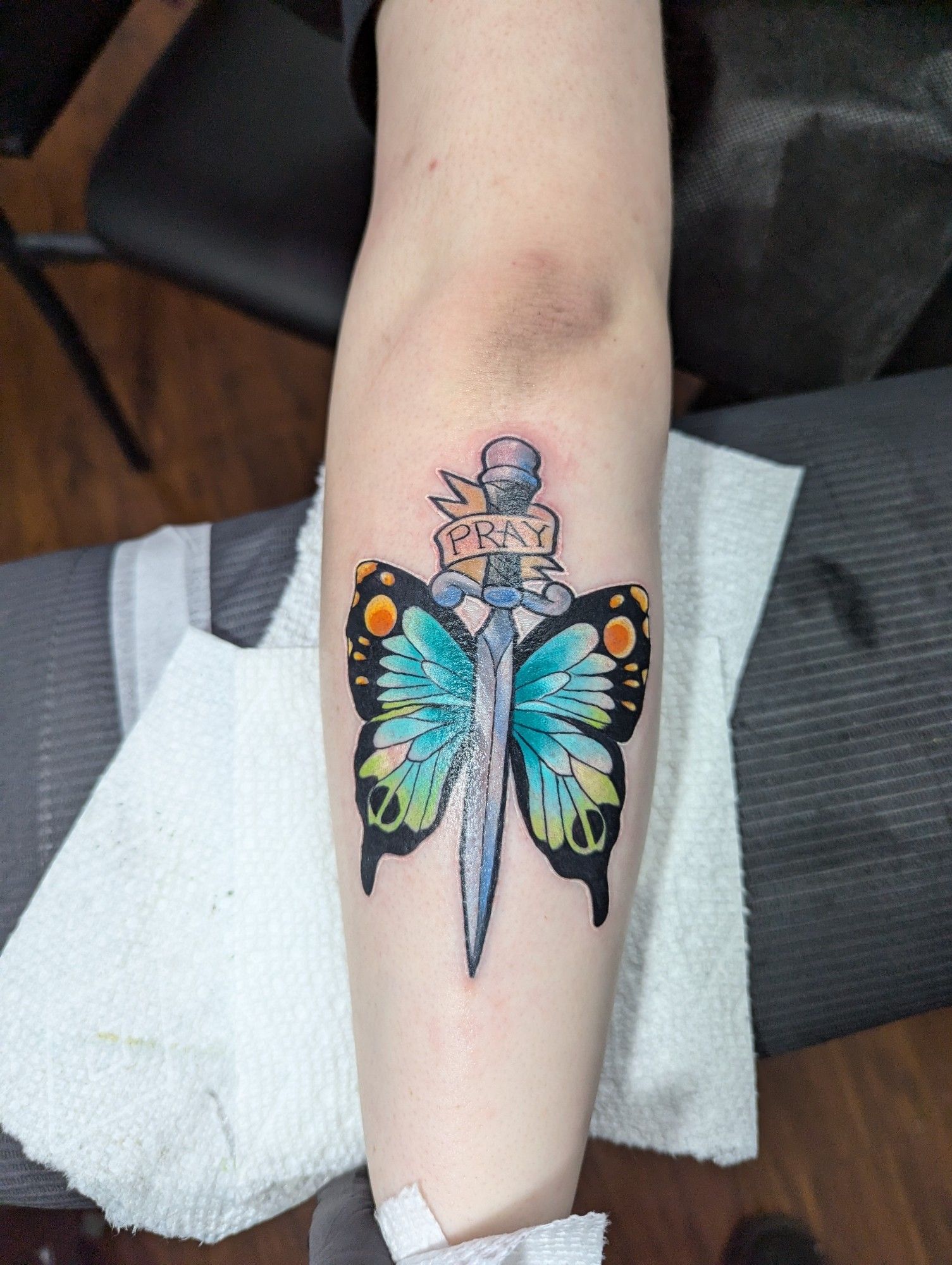 the back of lowercase judy's arm, now adorned with a tattoo of a dagger with blue, green, and orange butterfly wings. around the hilt of said dagger is a ribbon with the word "PRAY" on it.

it is jolyne cujoh's elbow tattoo from JoJo's bizarre adventure part 6