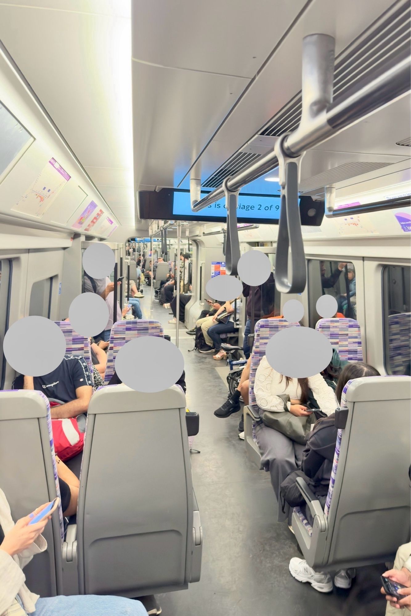 On a train running on the Elizabeth Line in London, showing a bright and open interior carriage with a good number of seated passengers and an overhead board partly obstructed by handrails.