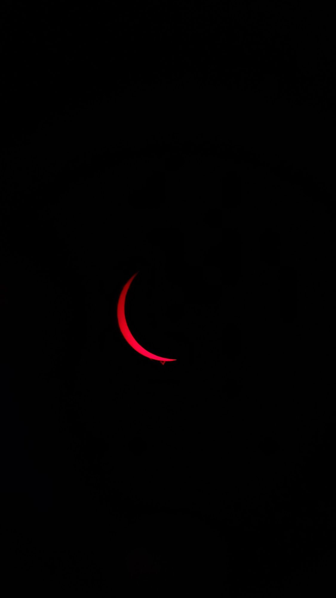 A blood red, very thin solar crescent in H-alpha lens, taken with a cellphone. A prominence at the lower edge of the sun.