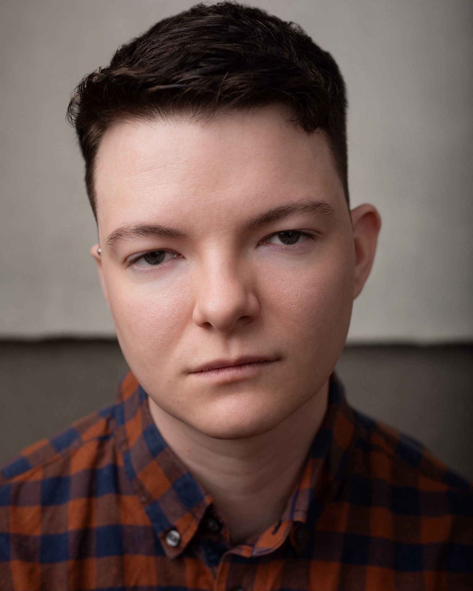 A young white man with short, dark hair in a tight fade faces the camera with a serious, intense expression. He is wearing a blue and reddish-orange plaid shirt with the top collar button undone.