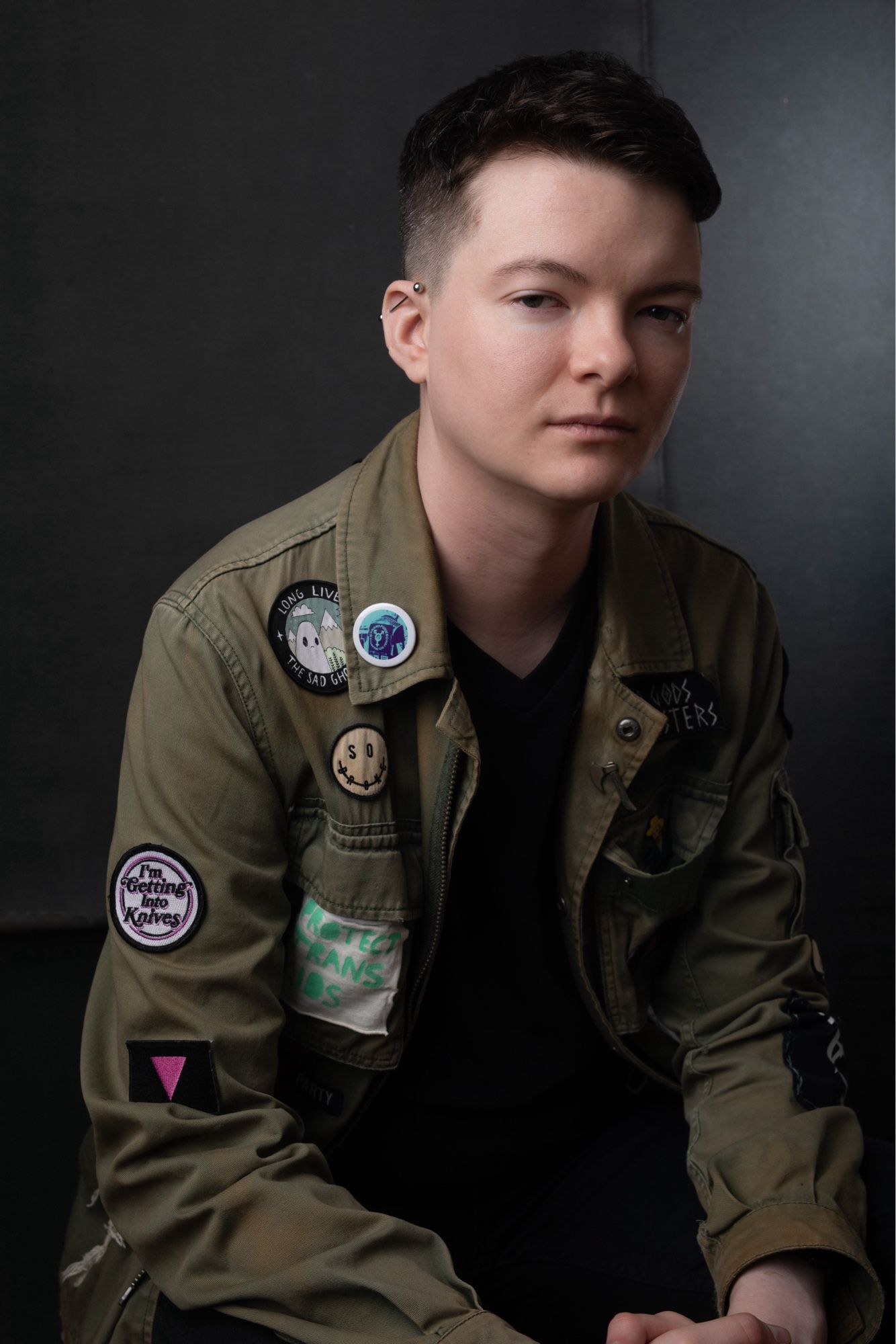 A young white man with short, dark hair in a tight fade in a seated pose in semi-profile, leaning slightly towards the camera. He wears a military-green patch jacket covered in patches with queer, anarchy, Satan, band, and other found art patches.