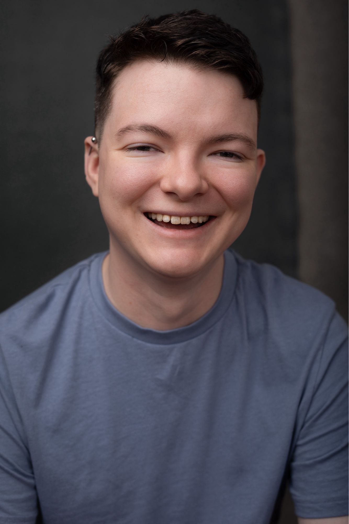 A young white man with short, dark hair in a tight fade faces the camera smiling, as though caught mid-laugh. He is wearing a light blue t-shirt.