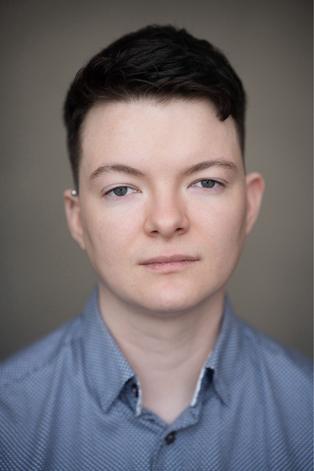 A young white man with short, dark hair in a tight fade faces the camera with a thoughtful, fairly neutral expression. He is wearing a light blue button shirt with the collar button undone.