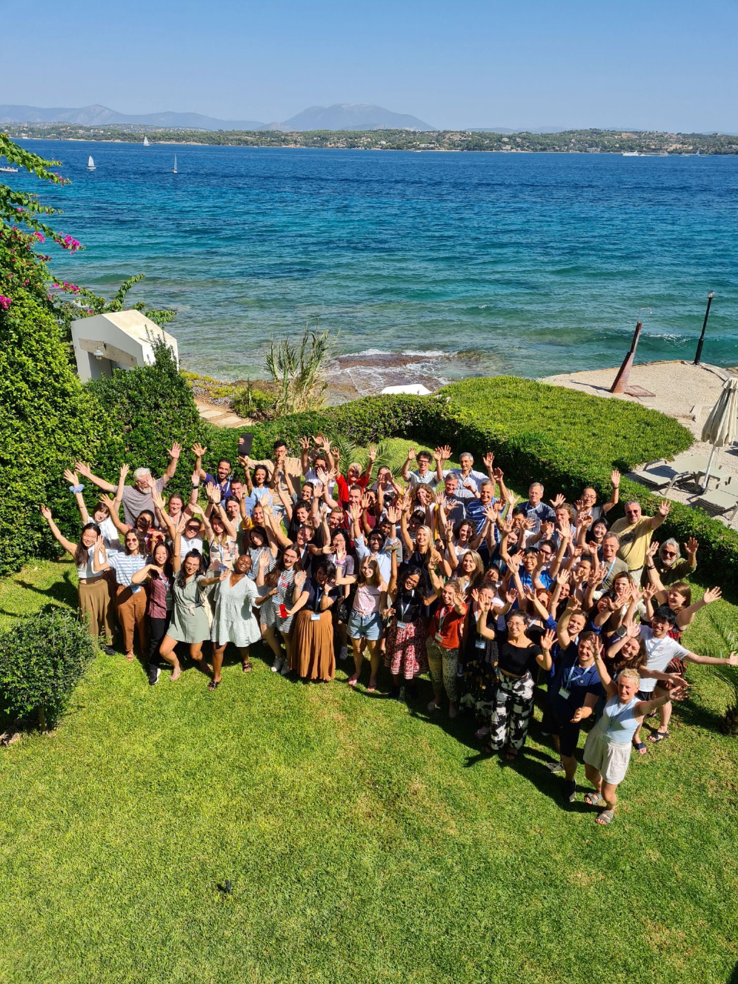 The delegates and faculty from the 2023 Summer School standing at the meeting location beside the beautiful Aegean sea, looking very happy.