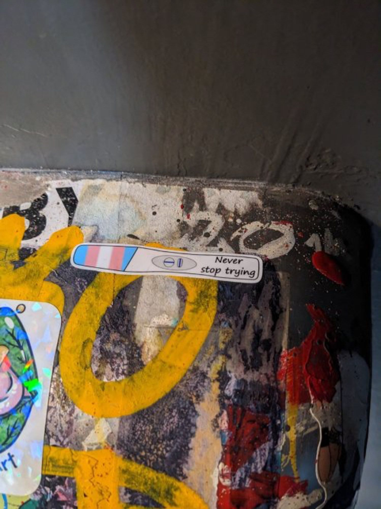 A sticker on something graffitied. On the left are vertical stripes in trans colors. On the right, the words 'Never stop trying'. It is a drawing of a pregnancy test