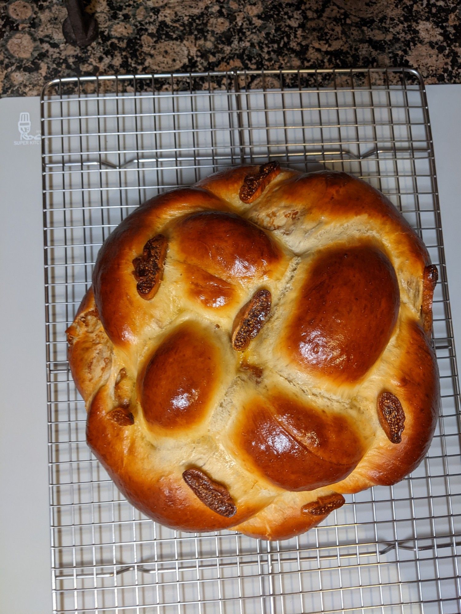 Golden and brown round fig challah cooling on a rack. The challah has a few sliced figs on top which have become jammy after being baked.