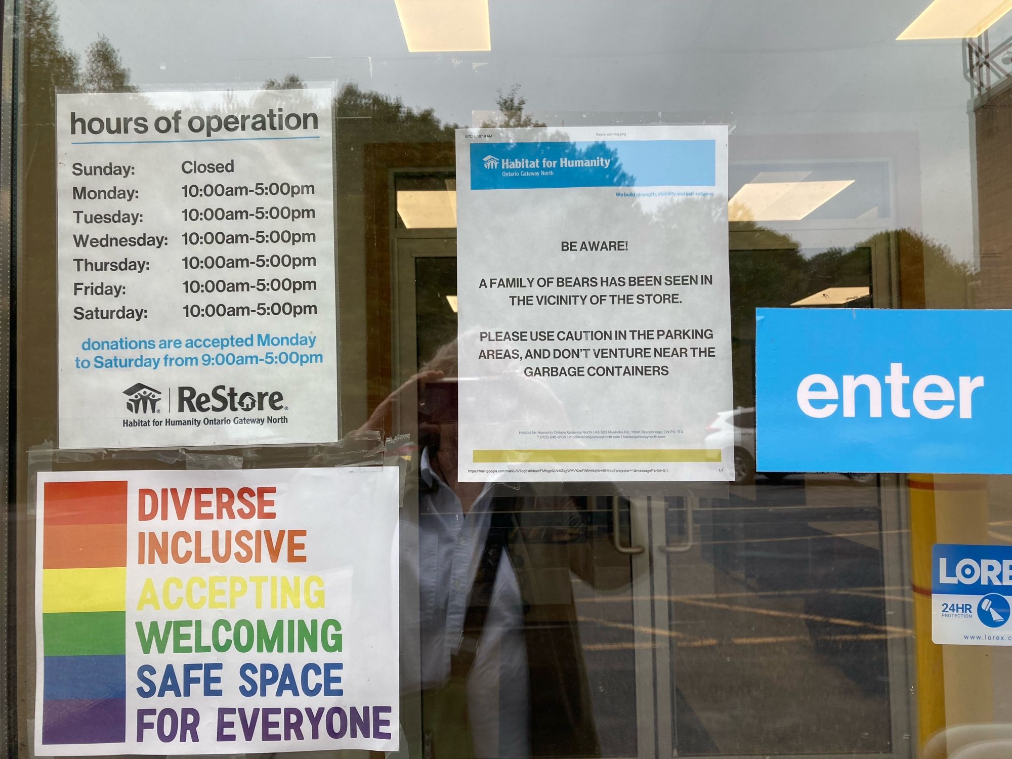 Signs on a door including a rainbow sign promoting tolerance and diversity, the store’s hours and a warning that there are bears in the vicinity.