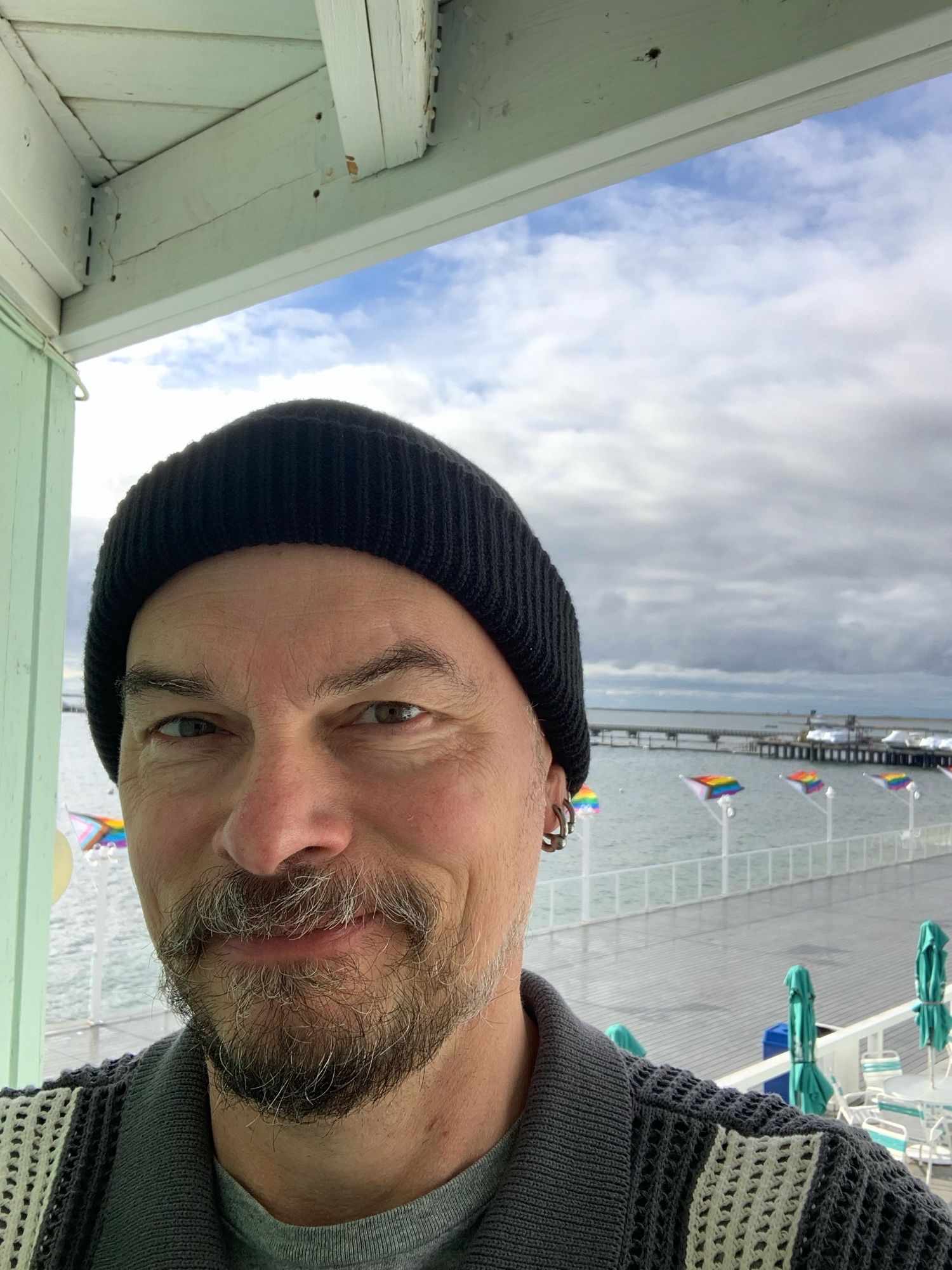 Steve on the balcony of his hotel room - in the background is cloudy skies, the water in the bay, queer flags blowing in the wind, a pier, and the deck of the hotel with tables and folded in umbrellas