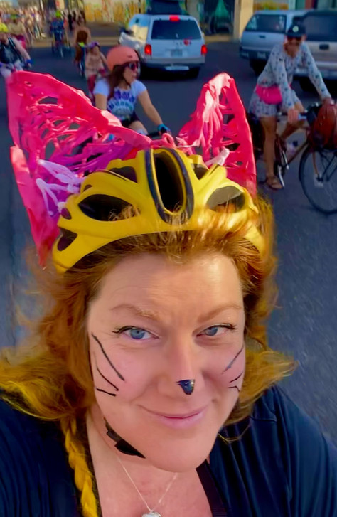me with a drawn on cat nose & whiskers, biking with yellow hair and a yellow helmet that I added oversized masking tape & pipe cleaner pink cat ears to
other bikers and big cars are behind me on a colorful street