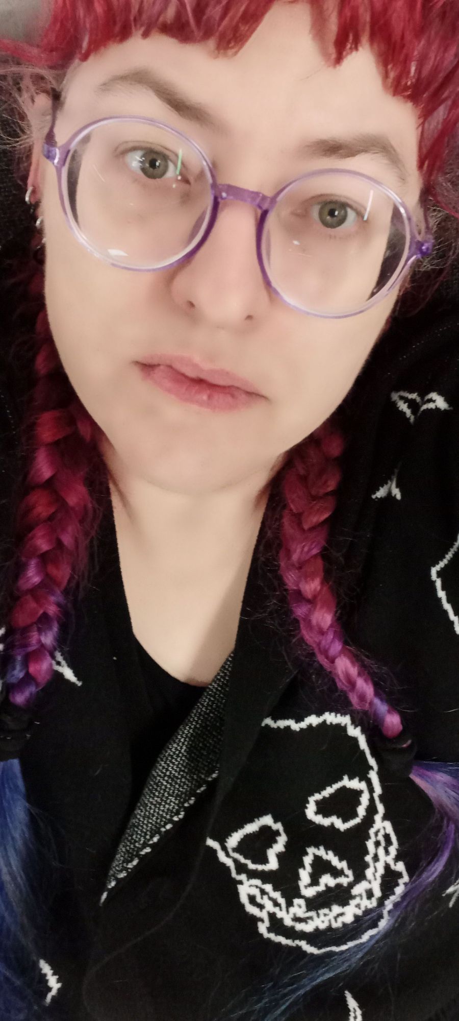 Female presenting person with purple hair in 2 braids and large glasses wearing a sweater with skulls on it