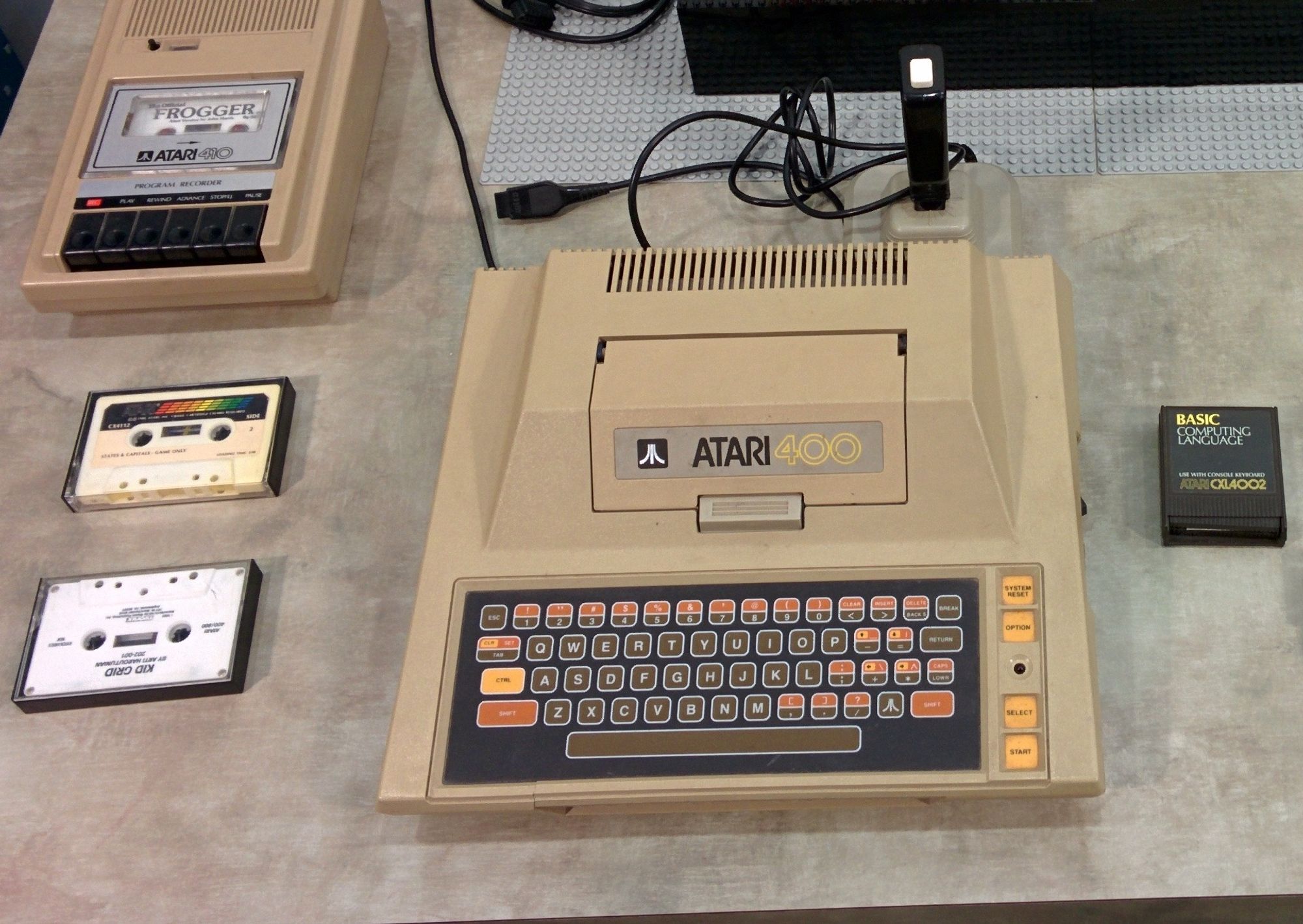 An Atari 400 home computer sits on a table. Several software cassette tapes sit near an Atari 410 tape drive. There's also a gray Wico Boss joystick and a BASIC language cartridge.