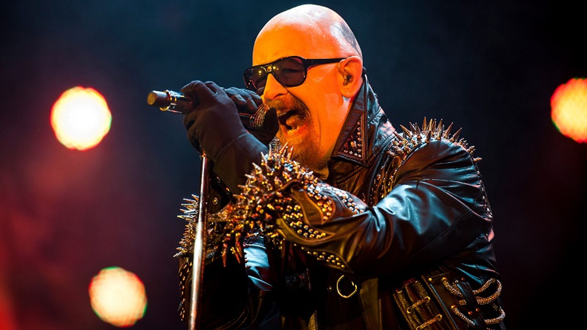 Rob Halford of Judas Priest, wearing shades and a black leather jacket covered in metal spikes, sings into a microphone on a stand.