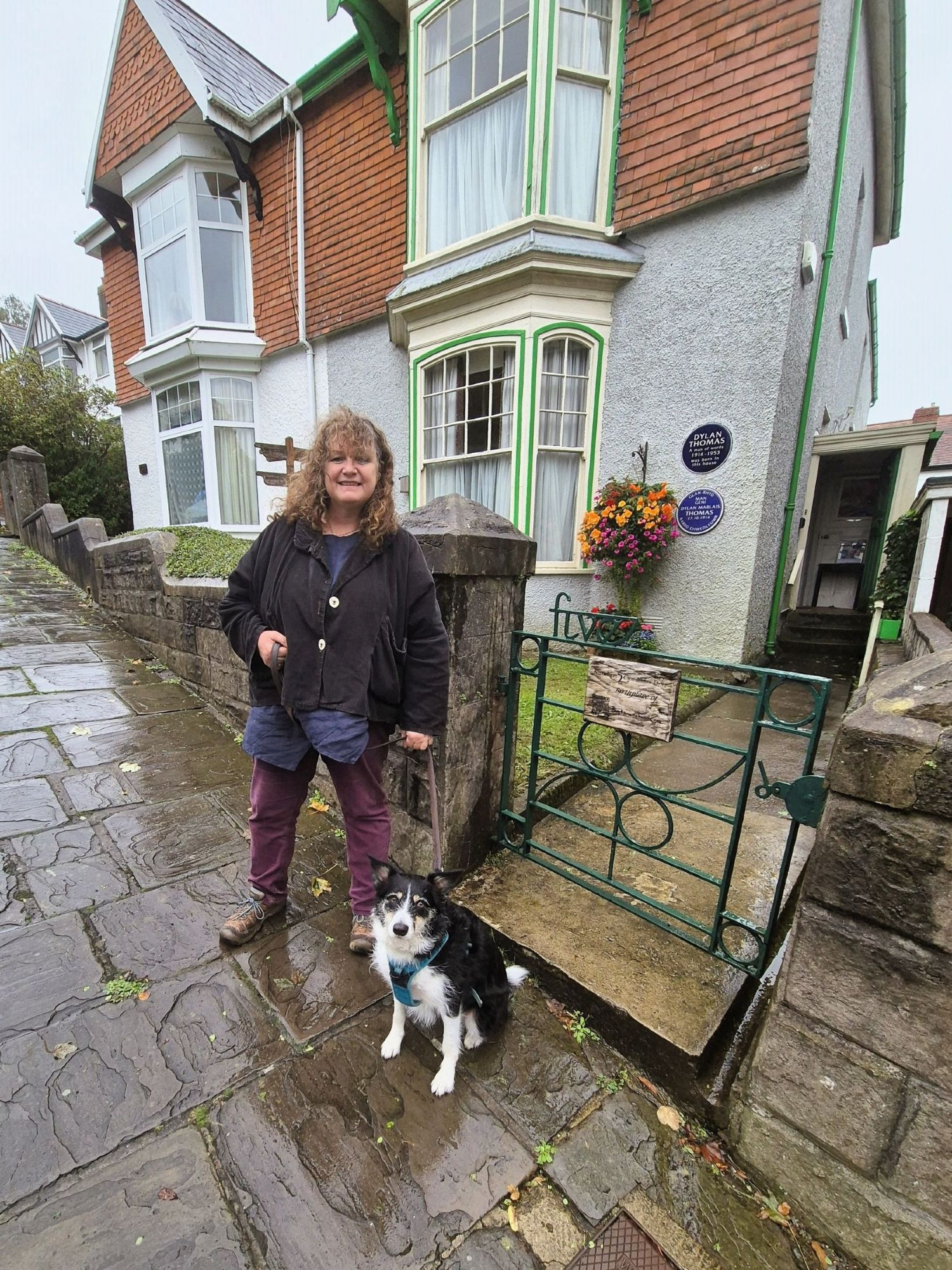 Woman and dog standing on rainy pavement outside Dylan Thomas's birthplace in Swansea, Wales. Two blue plaques on the wall behind - one in English, one in Welsh - and a hanging basket of petunias