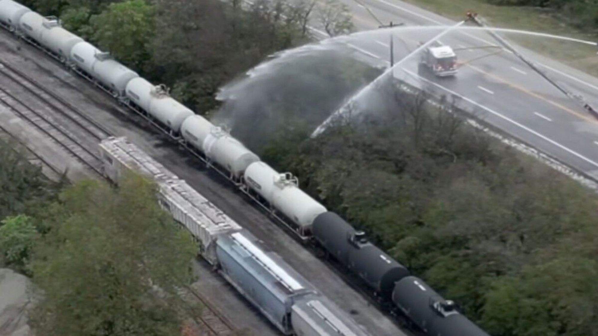 White railway tank cars leaking styrene, a toxic chemical. Two fire engines are spraying water on the tank cars from a long distance in an attempt to keep styrene vapors from escaping. Source: AP News