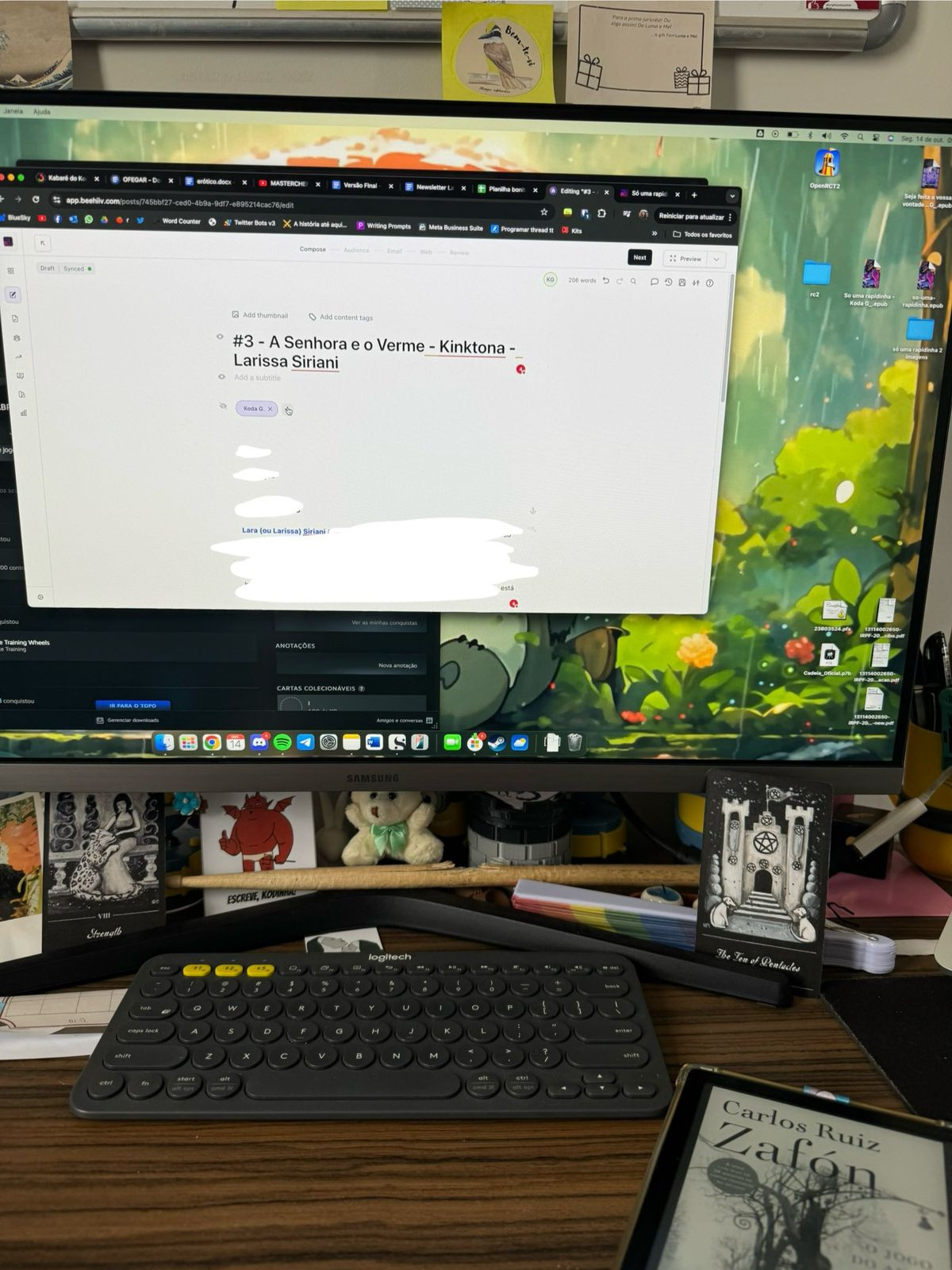 uma foto do meu monitor e teclado. no monitor, dá pra ver um site aberto e está escrito "#3 - a senhora e o verme - kinktona - larissa siriani". embaixo do monitor, um monte de coisinha minha, adesivos, cartas de tarot, uma baqueta, etc.