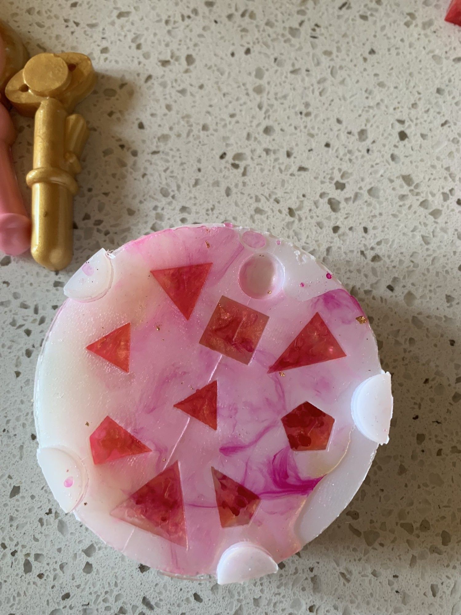 A photograph of a dice mold with resin dice curing inside - transparent pink and gold dice with the classic set of D20, a crystal d4, pyramid d4, d6, d8, 2 d10s, and d12.