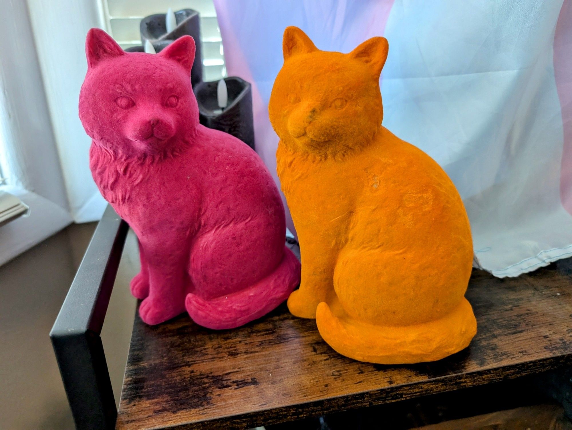 Two cat sculptures, one pink, one orange sit together on top of a shelf unit