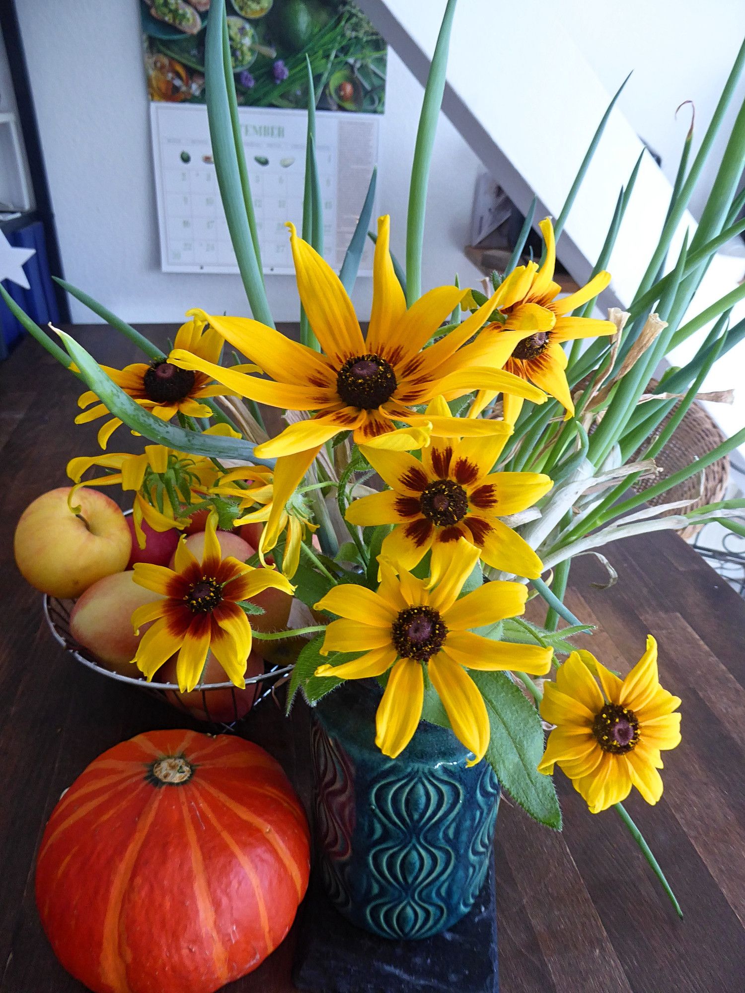 Blue vase with bright yellow, dark-centred Rudbeckias "Sahara". There's a bright orange Hokkaido pumpkin to the left of the vase. Behind it you can see a basket filled with apples and scallion shoots.