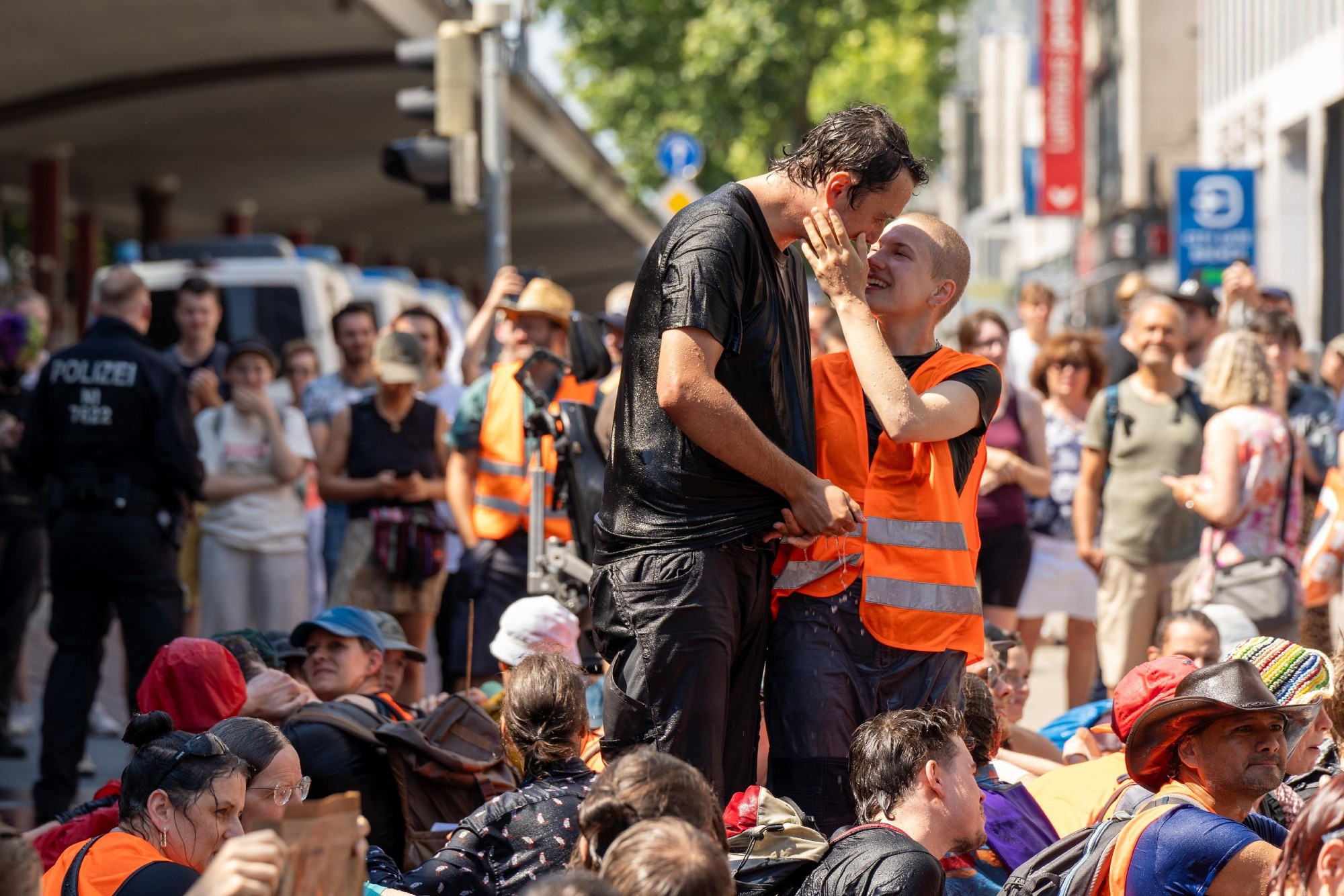 Eine Demonstration der Letzten Generation auf einer Straße bei sonnigem Wetter. Sie alle sind klatschnass von Wasserwerfern, sitzen jedoch friedlich zusammen. Inmitten von Ihnen stehen zwei Personen. Eine wringt sich gerade das Hemd aus. Die andere hat die Hand an der Wange der anderen Person und lächelt ihr zu. Ihre Gesichter sind nur wenige cm voneinander entfernt. Sie wirken sehr liebevoll und glücklich.