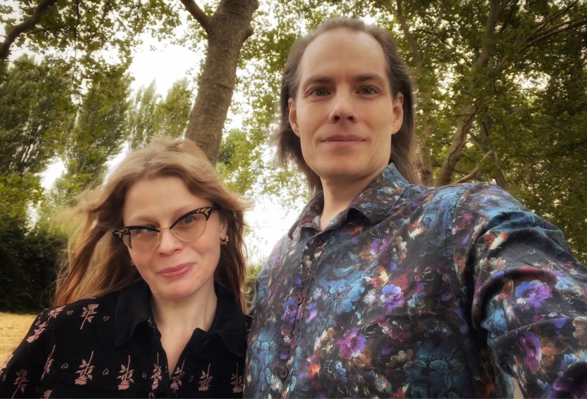 Two white people in mid-shot photo standing next to each other smiling for the camera, trees behind in a green space, woman on the left and a man on the right.