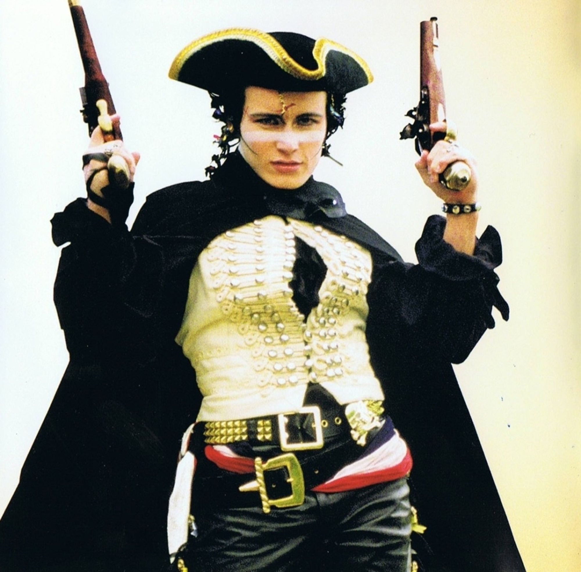 Adam Ant striking a pose with white face paint, a tricorn hat, two pistols, a cloak, a fancy ivory vest, two thick belts, a red white and blue cummerbund, and black leather pants. This is from his music video for Stand and Deliver.