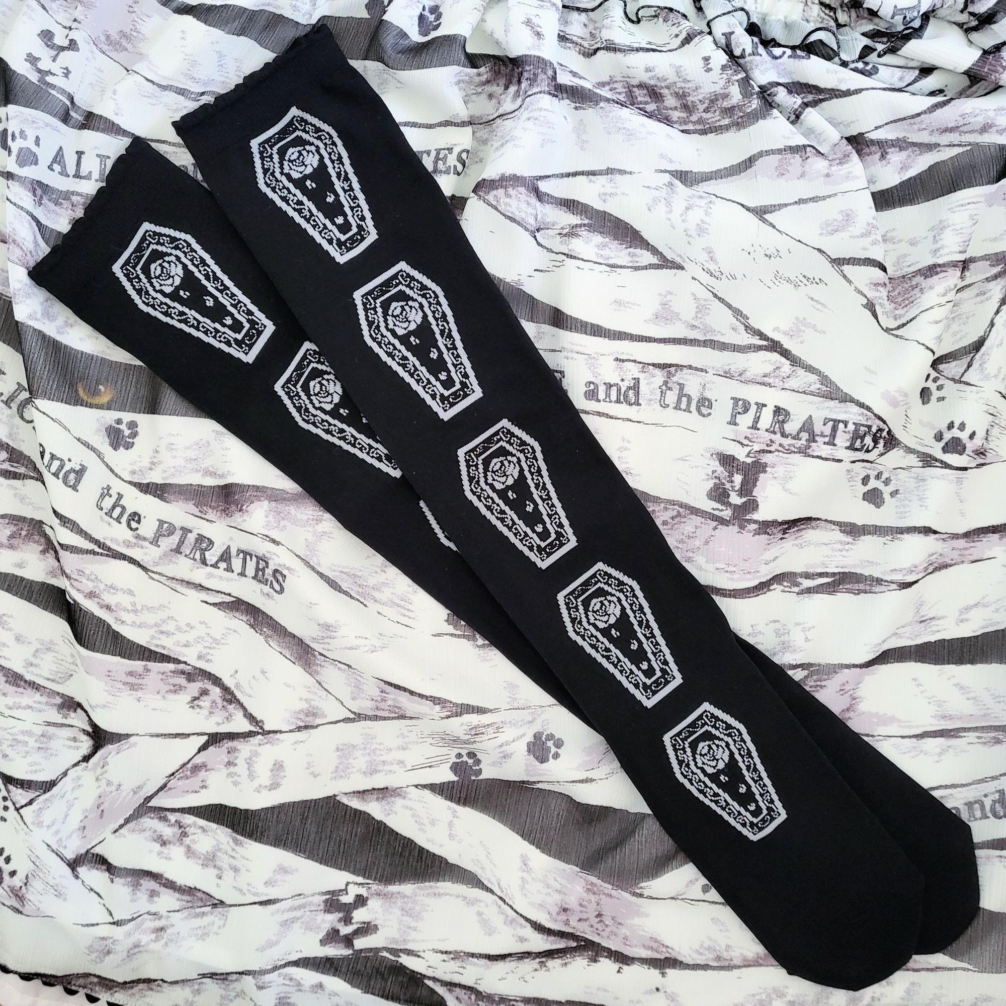 Black over knee socks laying down over a dress. The socks have coffins lined down the center. Each coffin has ornate filigree and a wilting rose.