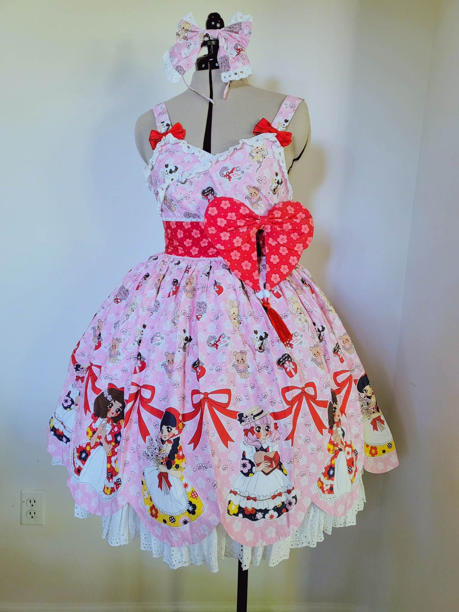 A pink lolita dress with flowers, bunnies, bears, and dogs all over it. It has a scalloped trim and 3 girls with ribbon along the border. The dress has red bows on the straps and a red waistband with large bow.