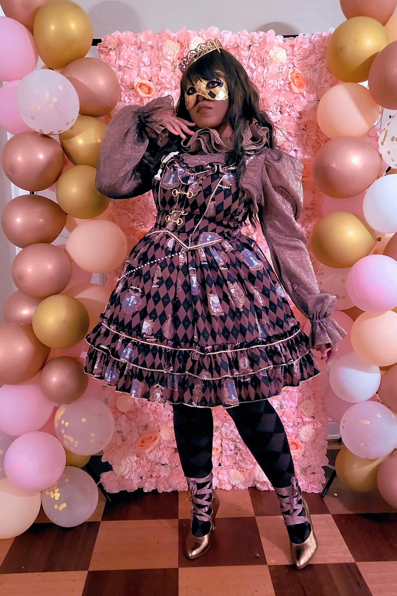 Milkcircus wearing a dark gold puffy blouse, brown x black harlequin patterned lolita dress, brown x black harlequin patterned socks, gold crown, gold mask, and gold boots.