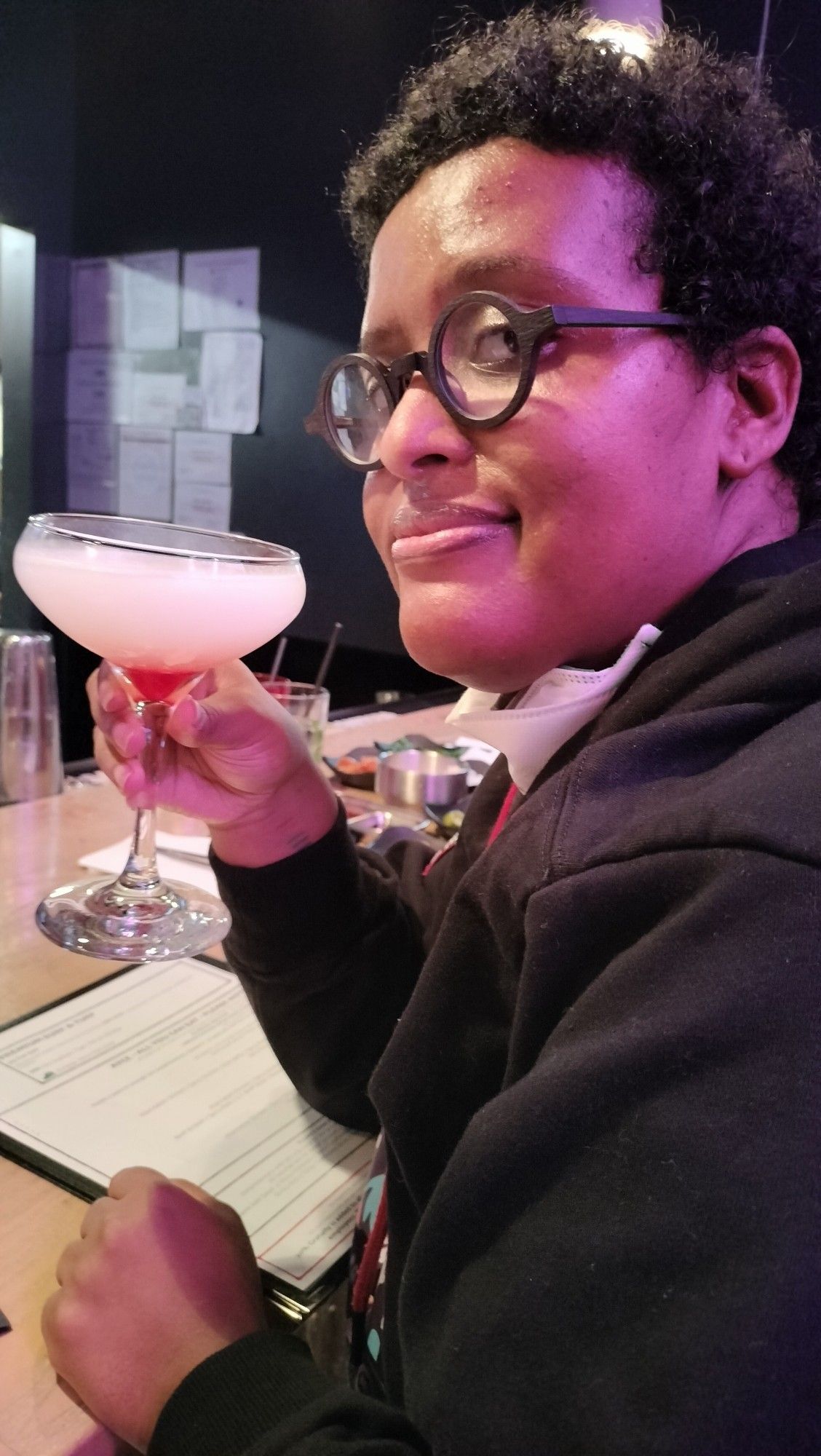 A picture of myself holding a cocktail. I am wearing glasses, a hoodie, and I am smiling