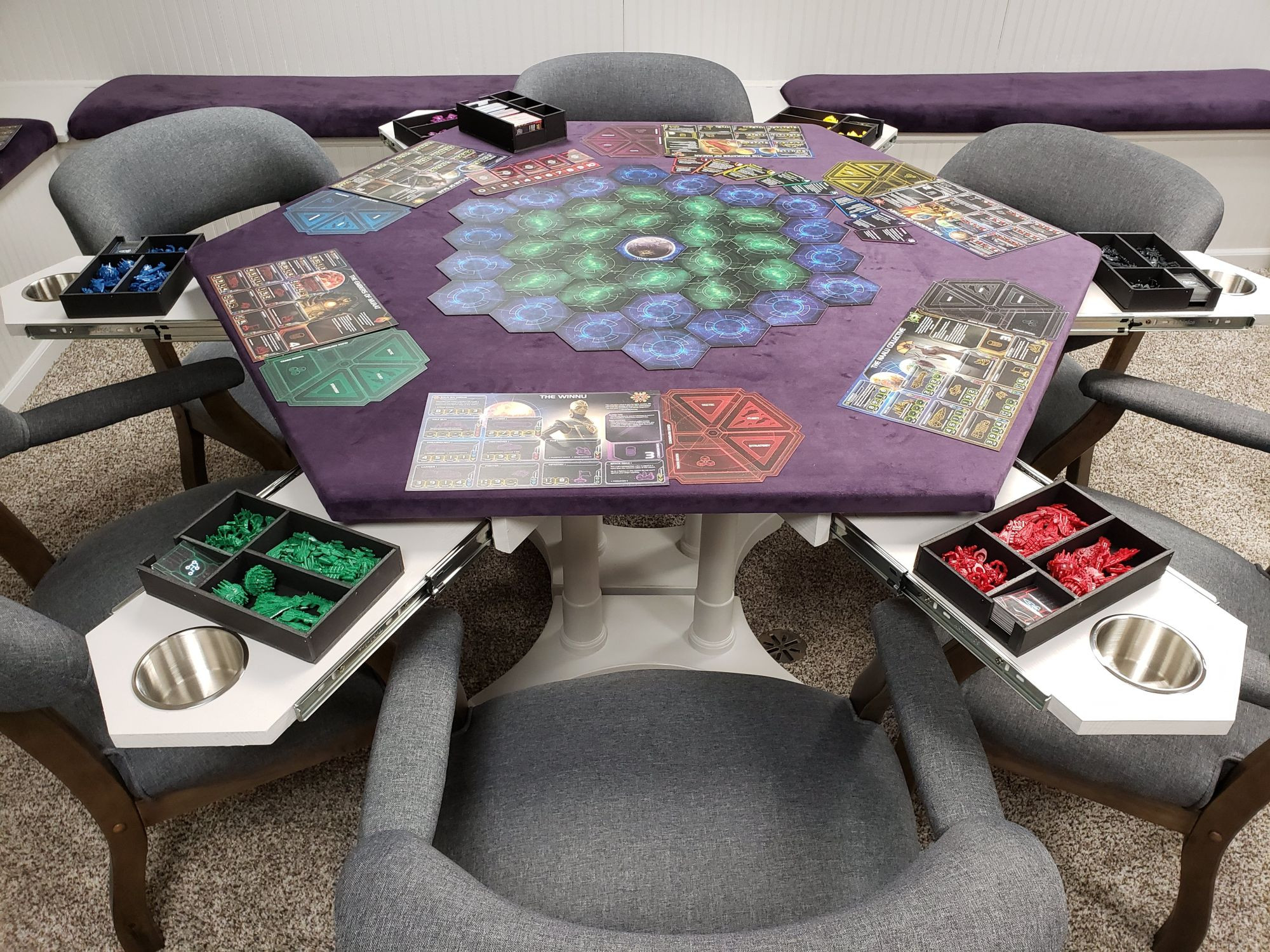 A purple hexagonal boardgame table surrounded by six chairs. There are slideout trays at each corner, with a drink holder in each. 
Setup on the table is the start of the boardgame Twilight Imperium 4.