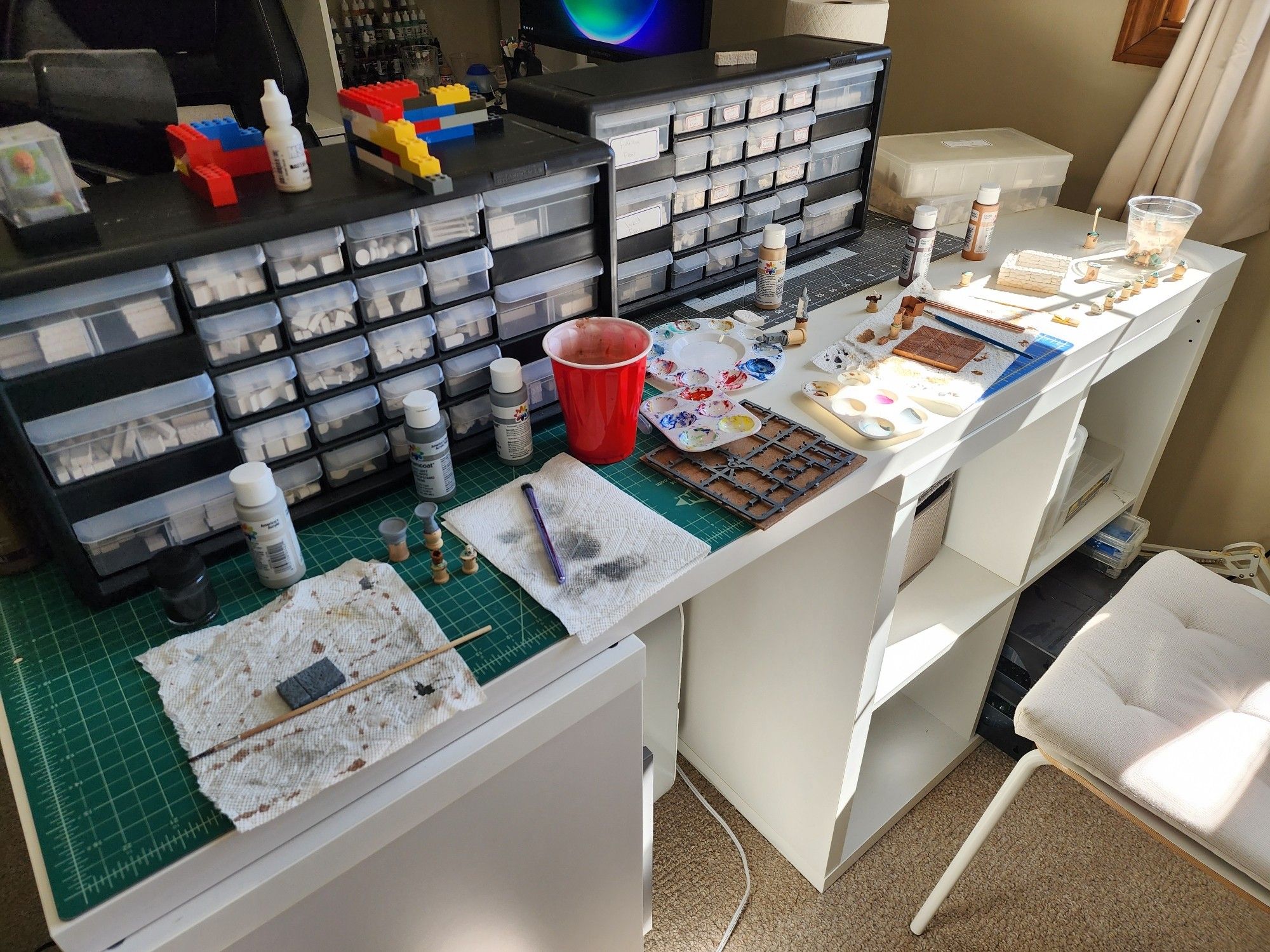 A hobby desk with cutting mats, paint brushes, paints and some bins of plaster pieces. It's a mess, with two painting stations to work at.