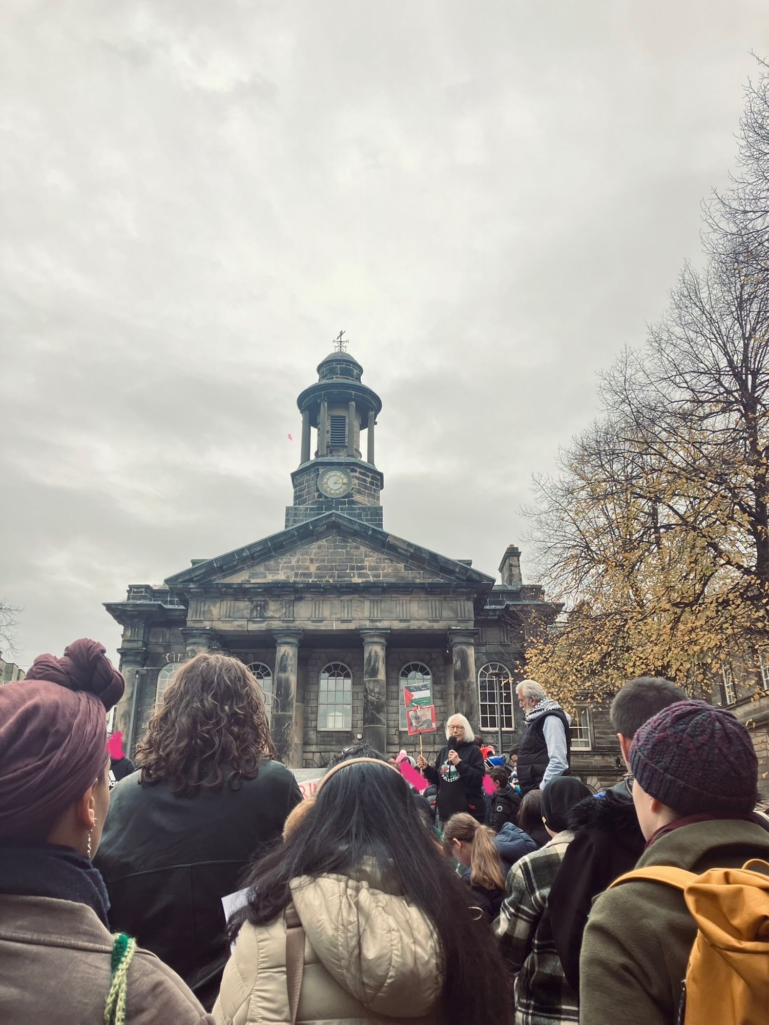 Backs of vigil attendees in a town square, looking toward a stage with a Jewish speaker and an Arab speaker standing together, on an overcast day.